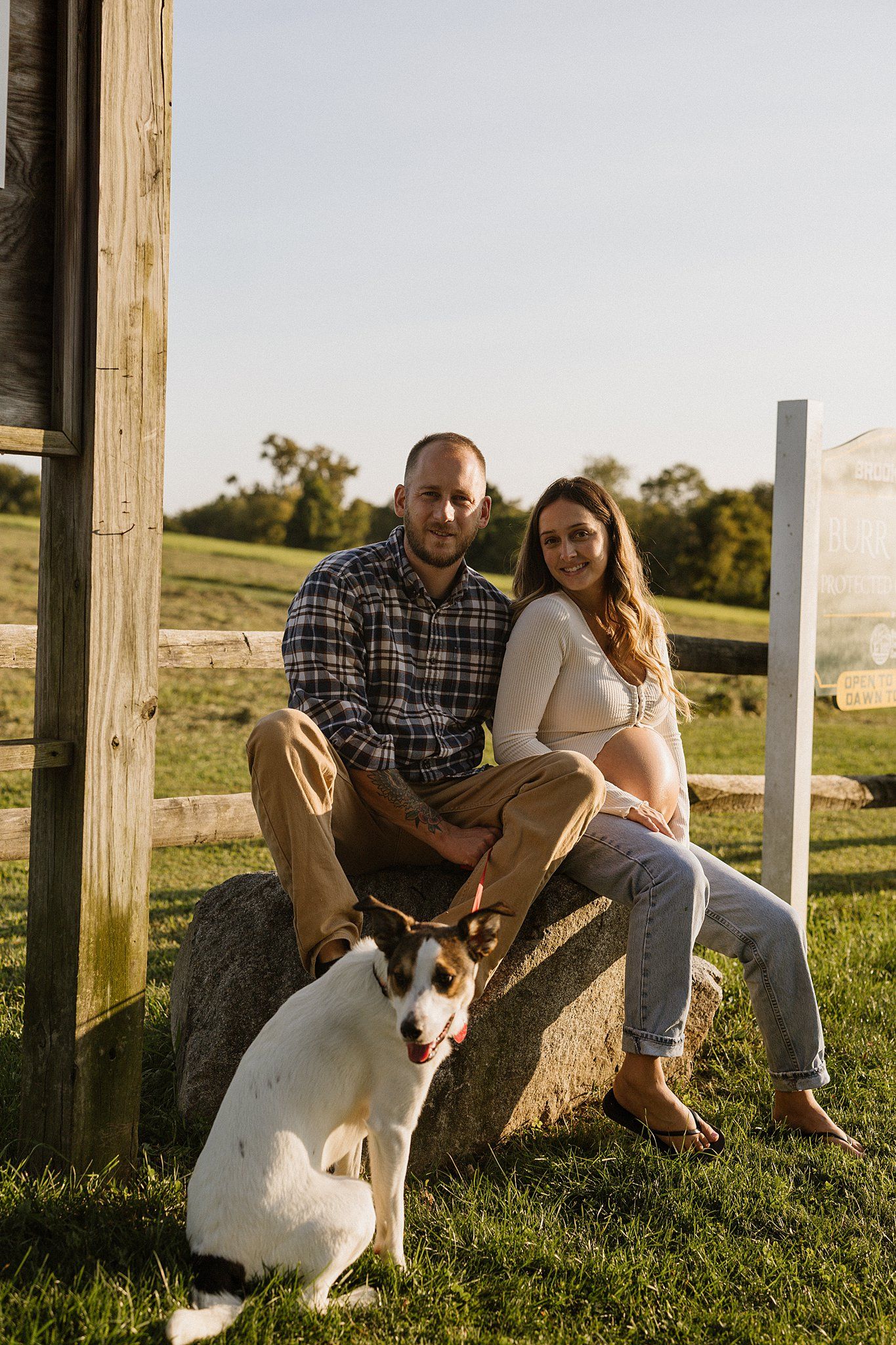 Couple sitting on fence post with their dog in countryside setting.