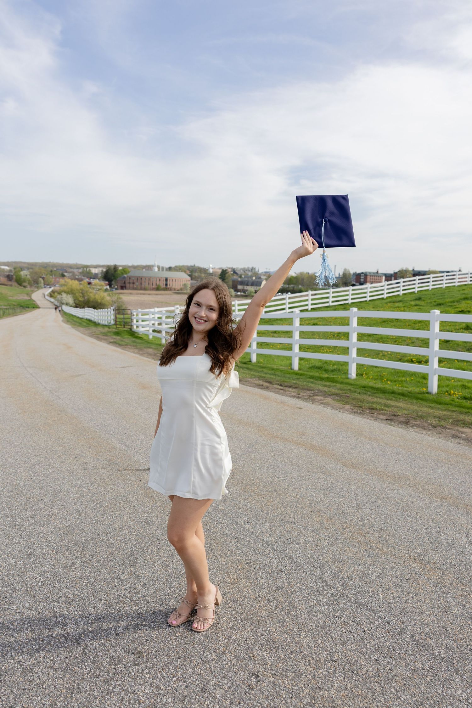 Graduate in white dress holds up blue cap while celebrating outdoors near white fence and rolling hills.