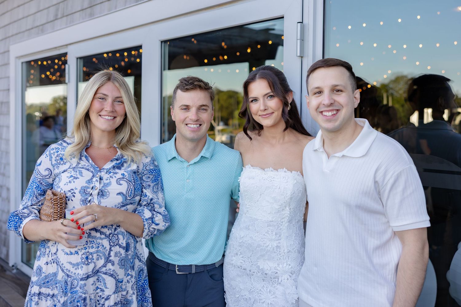 Wedding party poses outdoors against white building backdrop with string lights.
