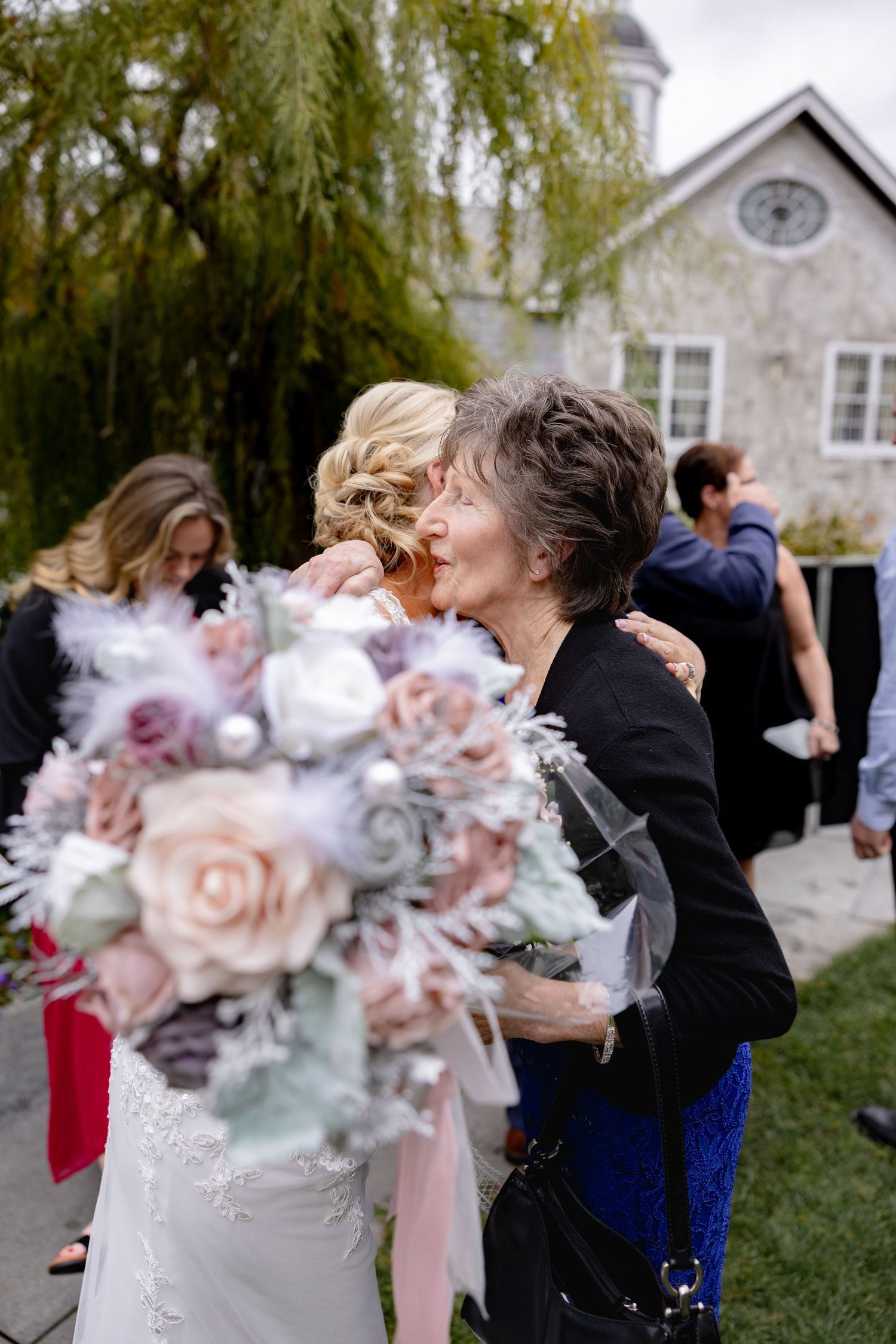 A touching moment at a wedding as guests embrace and admire a soft pink and gray floral bouquet.