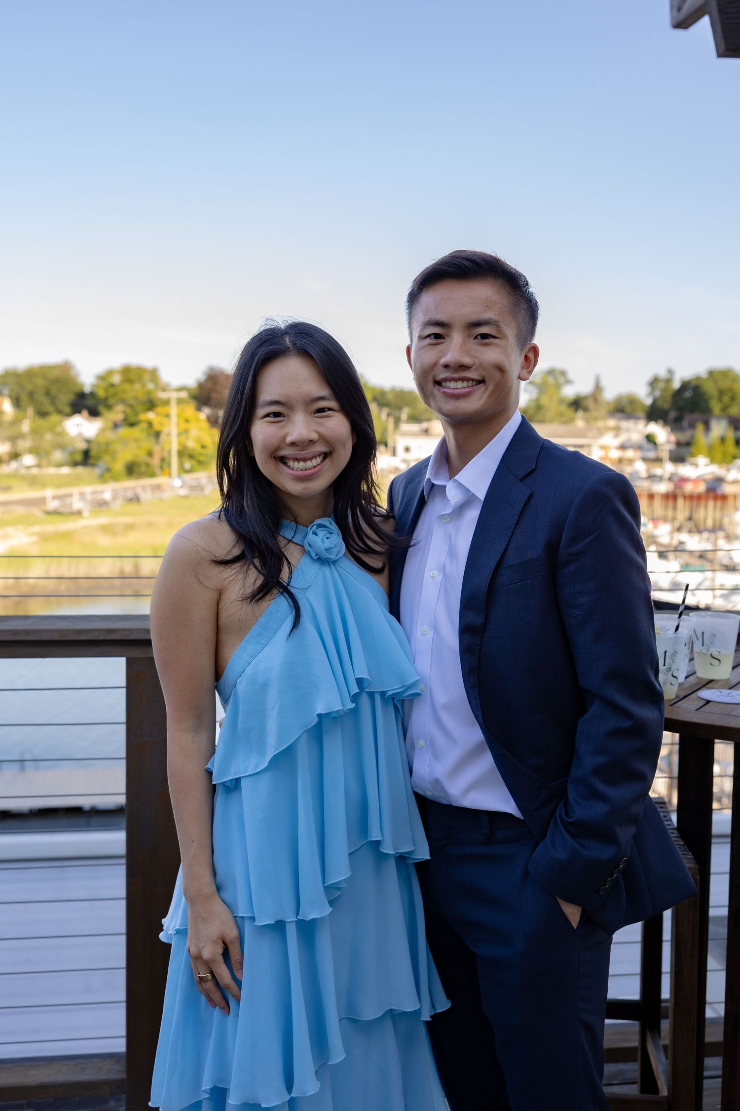 Couple posing together on a deck with a marina view, woman wearing light blue ruffle top.