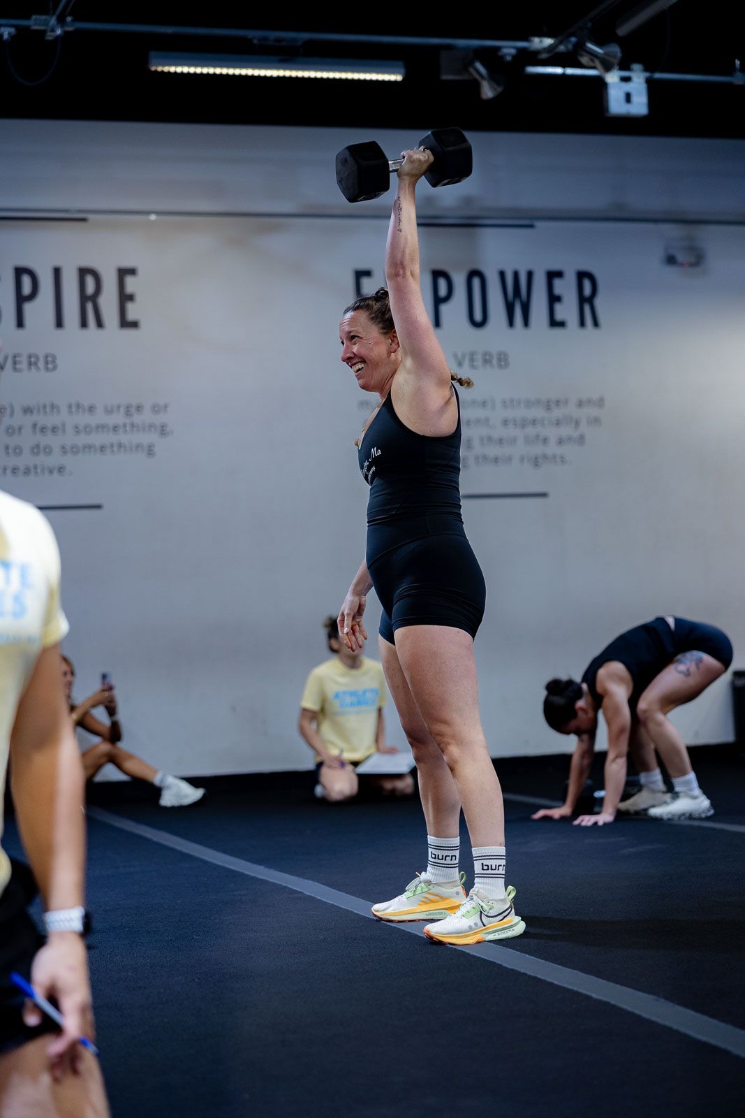 Person in black workout attire lifting a dumbbell overhead during a fitness class with motivational wall text.
