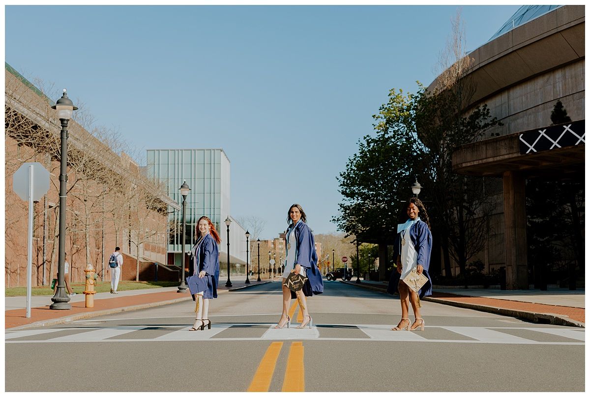 Three graduates in blue robes pose in the middle of a campus street.