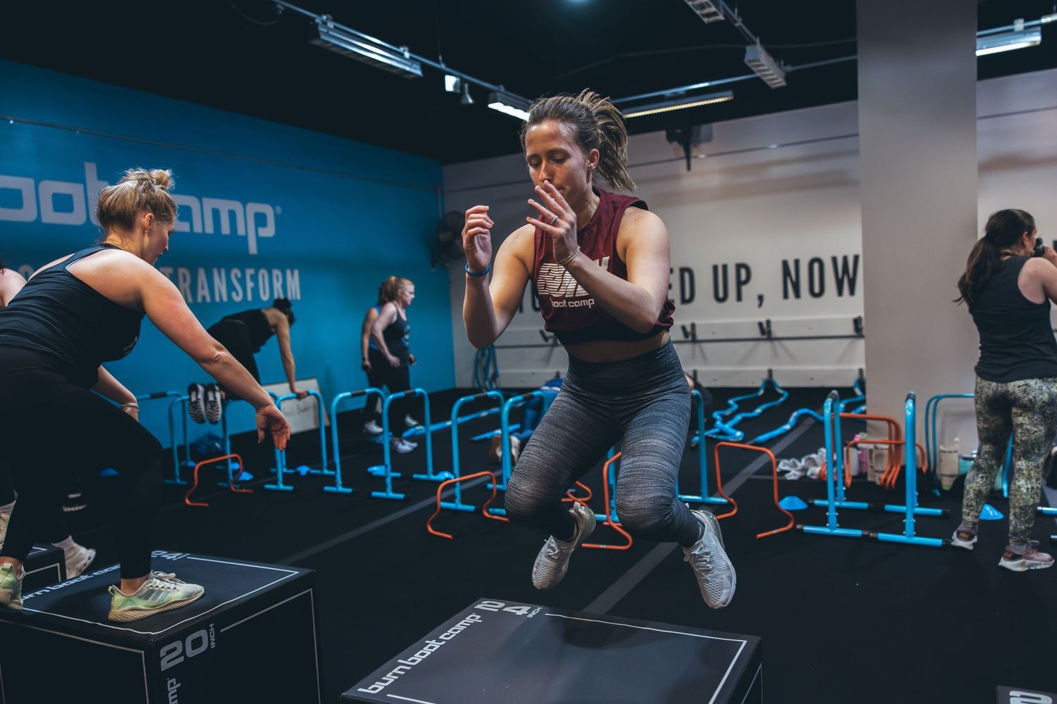 An athlete performs a dynamic jump exercise during an intense workout session in a blue-lit gym setting.
