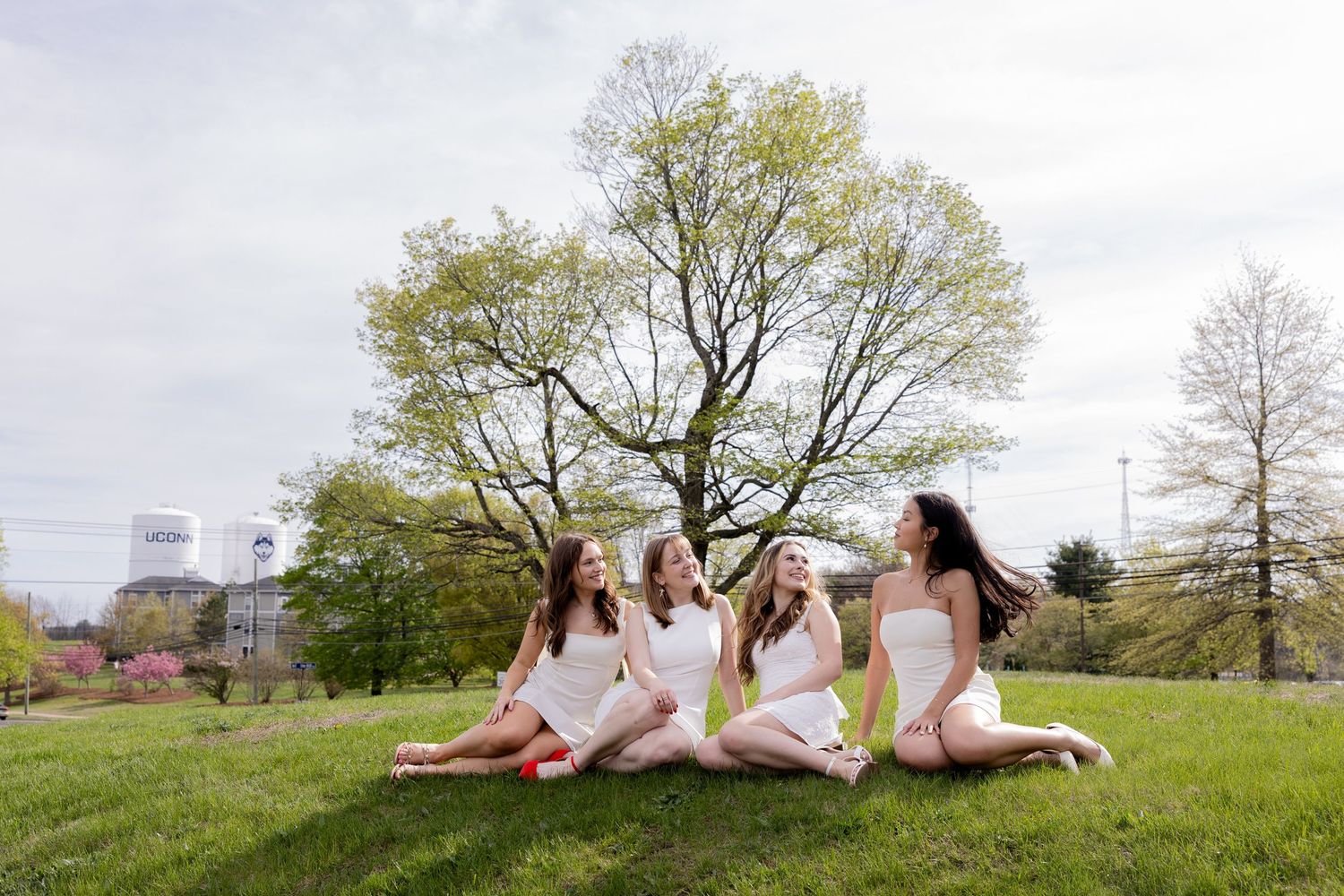 Group of friends in white dresses sitting together on a grassy hill during springtime with city buildings in background.