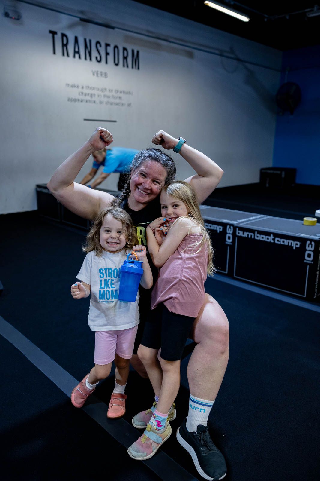 Three people pose together at Transform gym, flexing and celebrating in front of a blue illuminated wall.