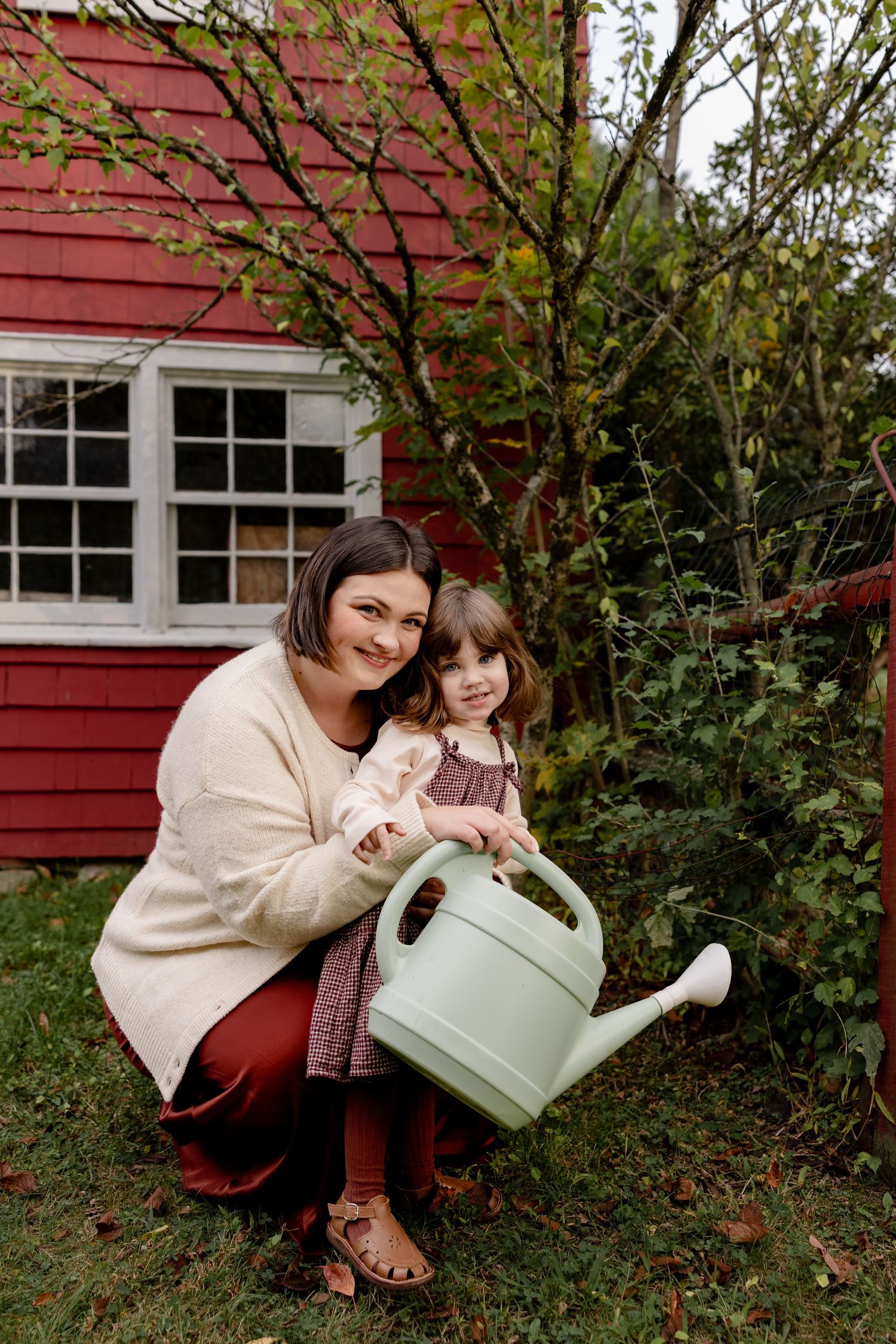 A pair of people gardening together with a mint green watering can beside a red barn in autumn.