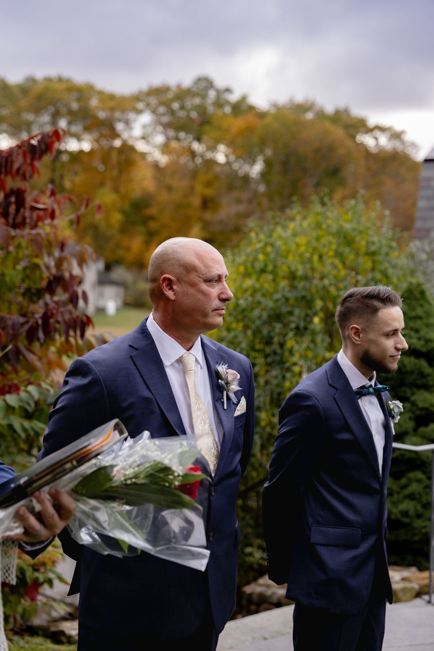 Two men in navy suits walk outdoors during autumn, one carrying a bouquet of flowers.