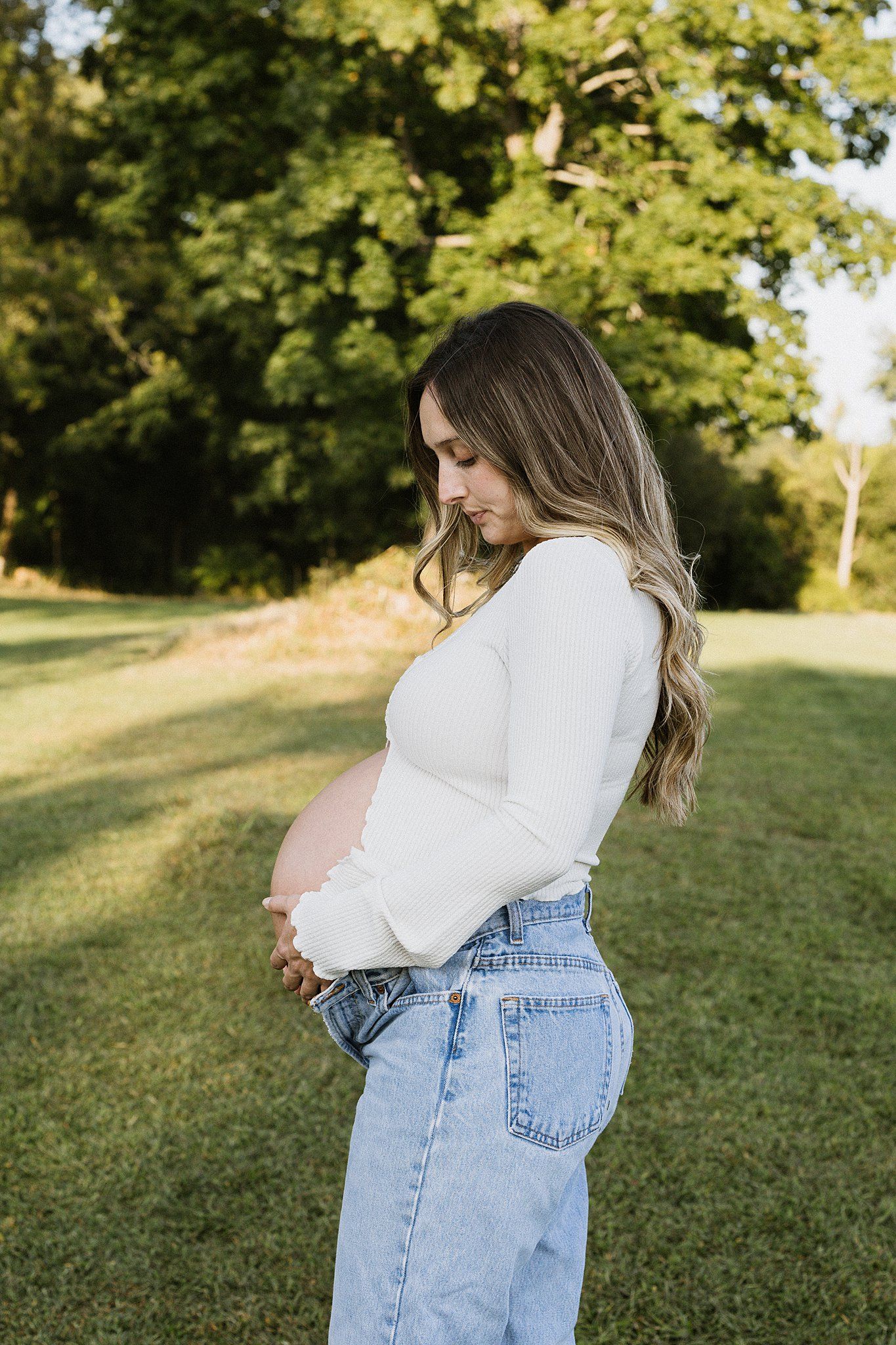 Side view of pregnant woman cradling belly in casual outfit against natural backdrop.