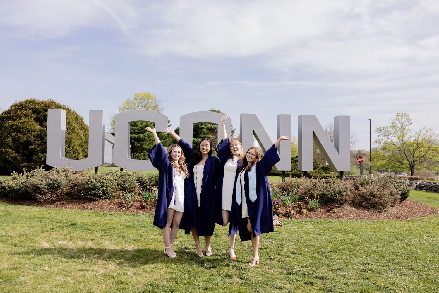Three graduates in blue caps and gowns celebrate by large UCONN letters outdoors on a grassy area.