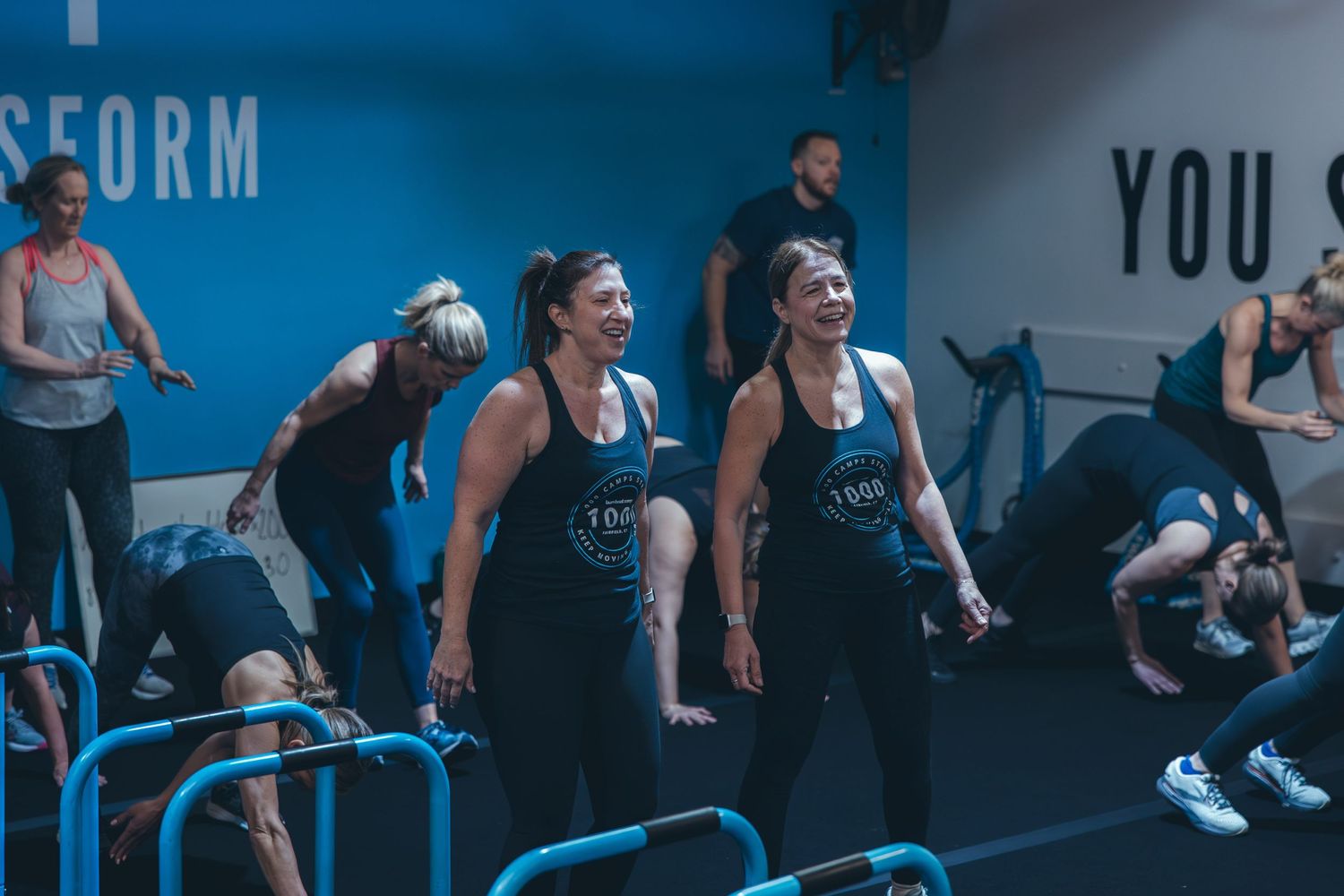 Group fitness class participants exercising together in a modern blue-lit gym space.