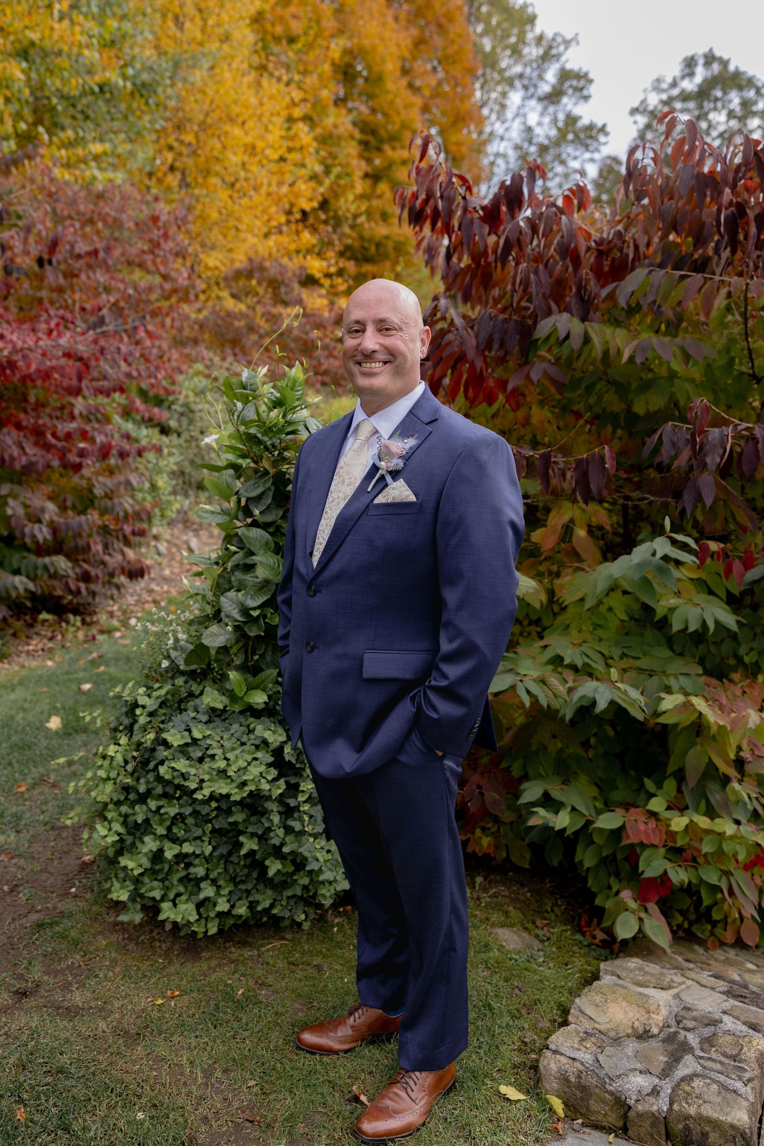 A groom in a navy suit poses outdoors against autumn foliage with vibrant red and yellow leaves.