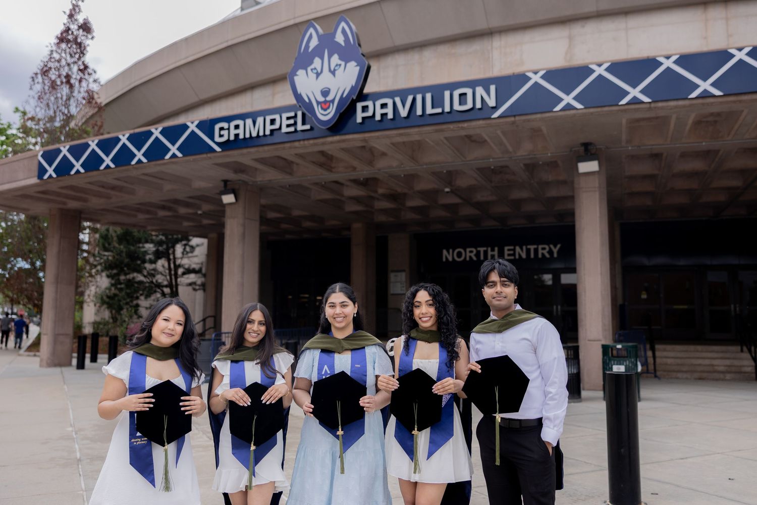 A group of graduates in blue academic regalia pose outside the Ramdel Pavilion entrance.