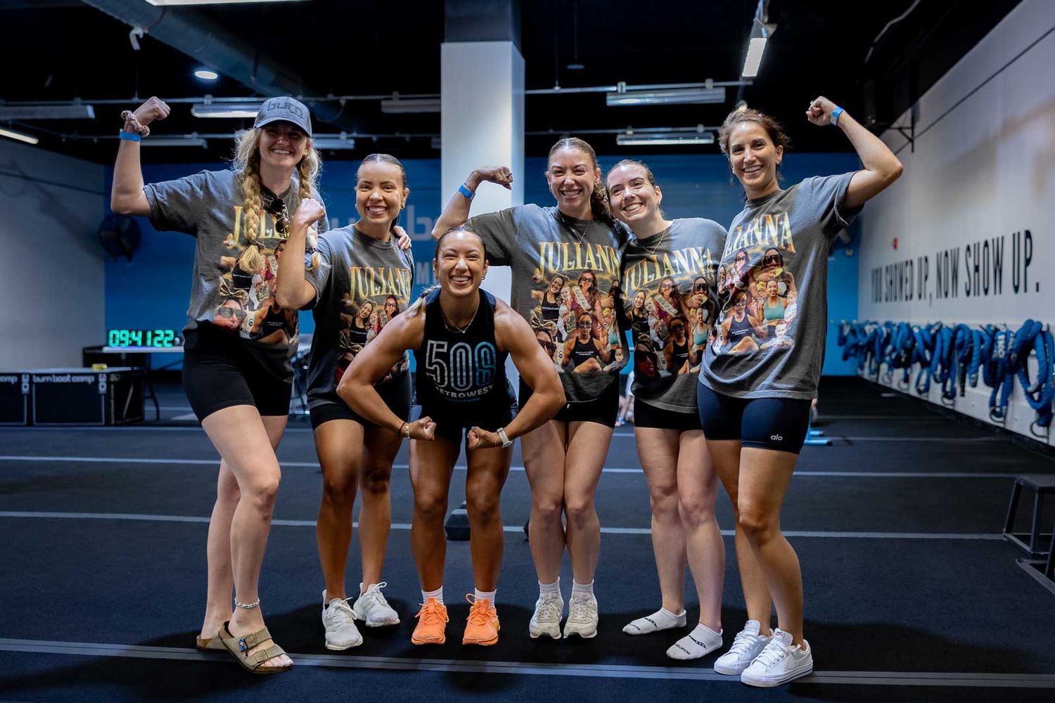 A group of athletes in workout attire pose together with thumbs up at a fitness gym.