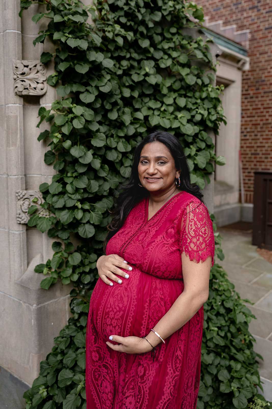 A person wearing a red lace dress poses outdoors against a brick wall with climbing ivy.