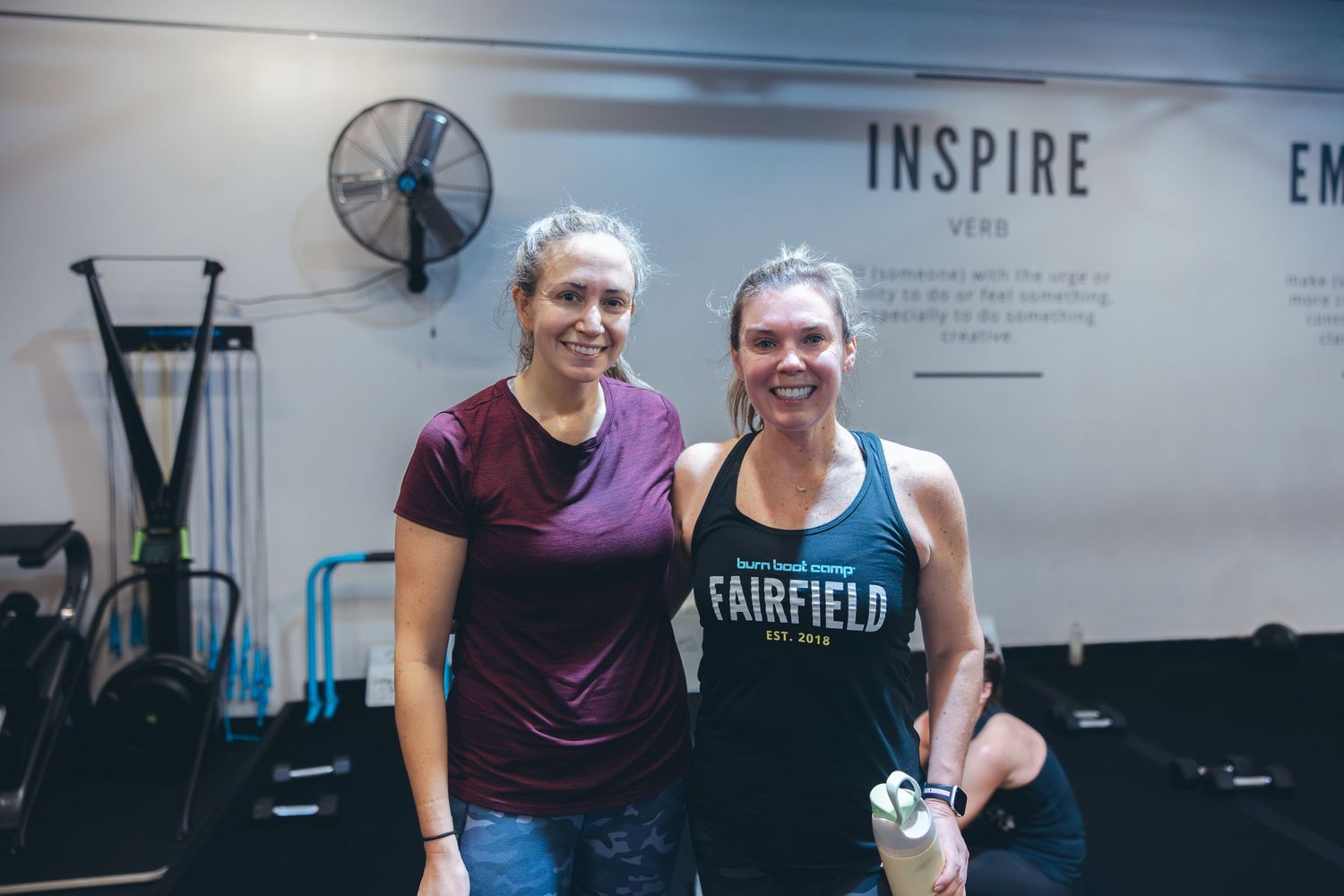 Two people in workout attire smiling in front of an 'Inspire' sign at a fitness studio.