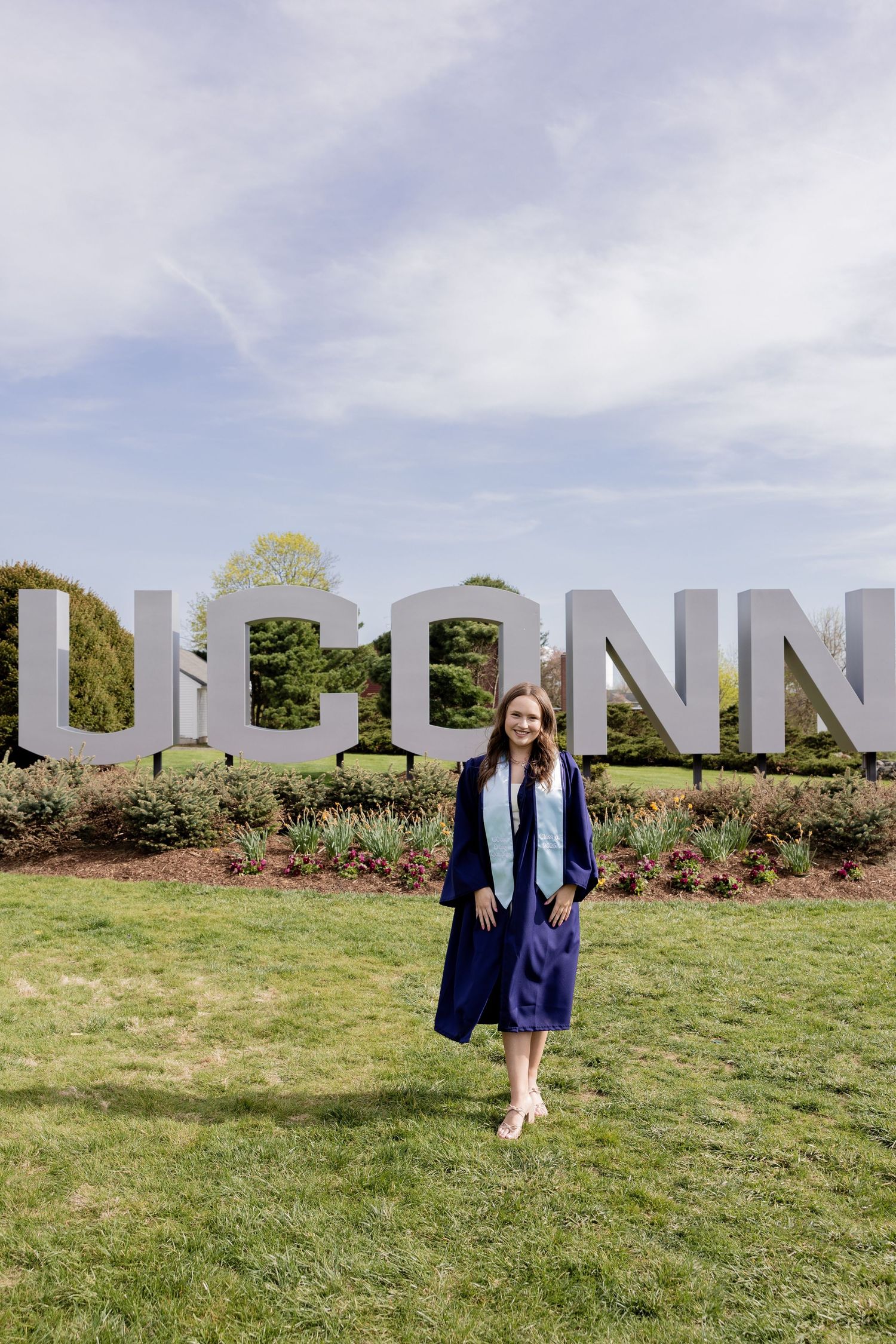 Graduate in blue cap and gown stands on grass in front of large UCONN letters on a sunny spring day.