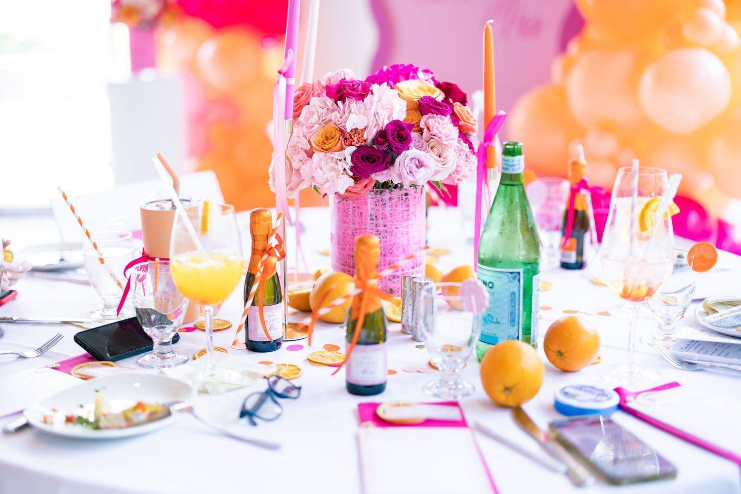 Colorful party table setting with pink and orange decorations, balloons, drinks and snacks in bright natural light.