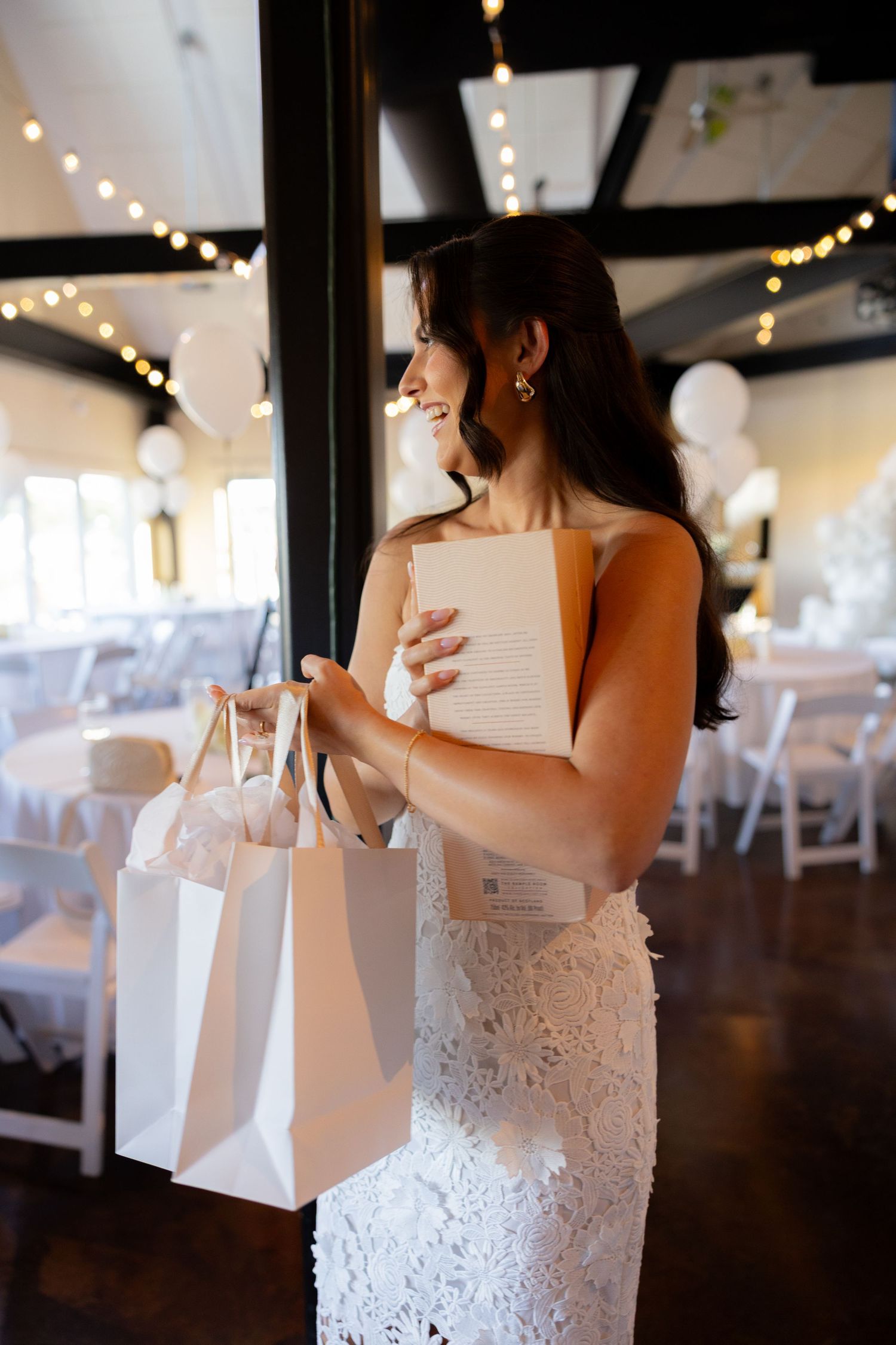 White gift bags are being held in a reception hall decorated with string lights and white chairs.