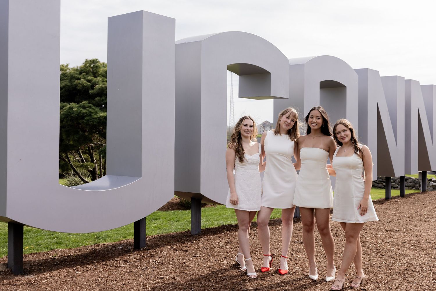 Four people in white dresses pose together in front of large white letters outdoors.