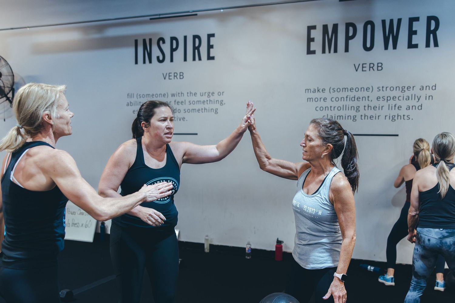 Three people high-fiving during a group workout session in front of an inspirational wall display.