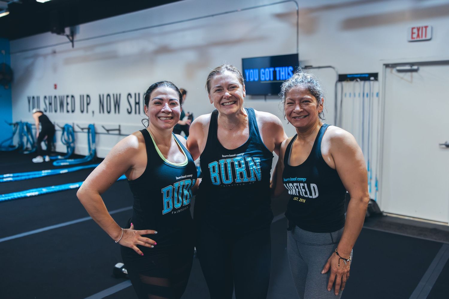 Three women in matching BURN tanks posing together at a fitness studio.