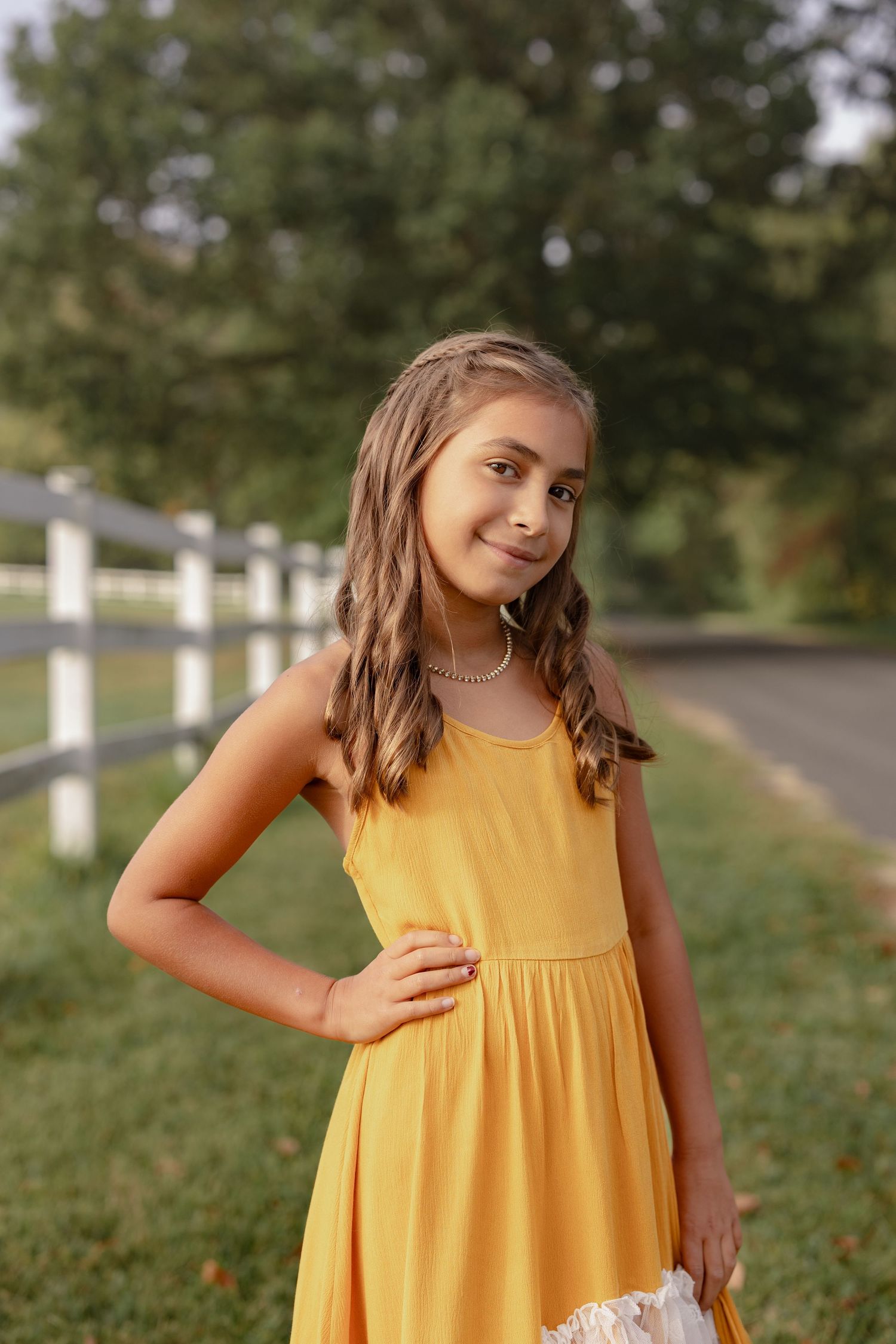 A young person in a bright yellow sundress poses outdoors on a grassy path lined with white posts.