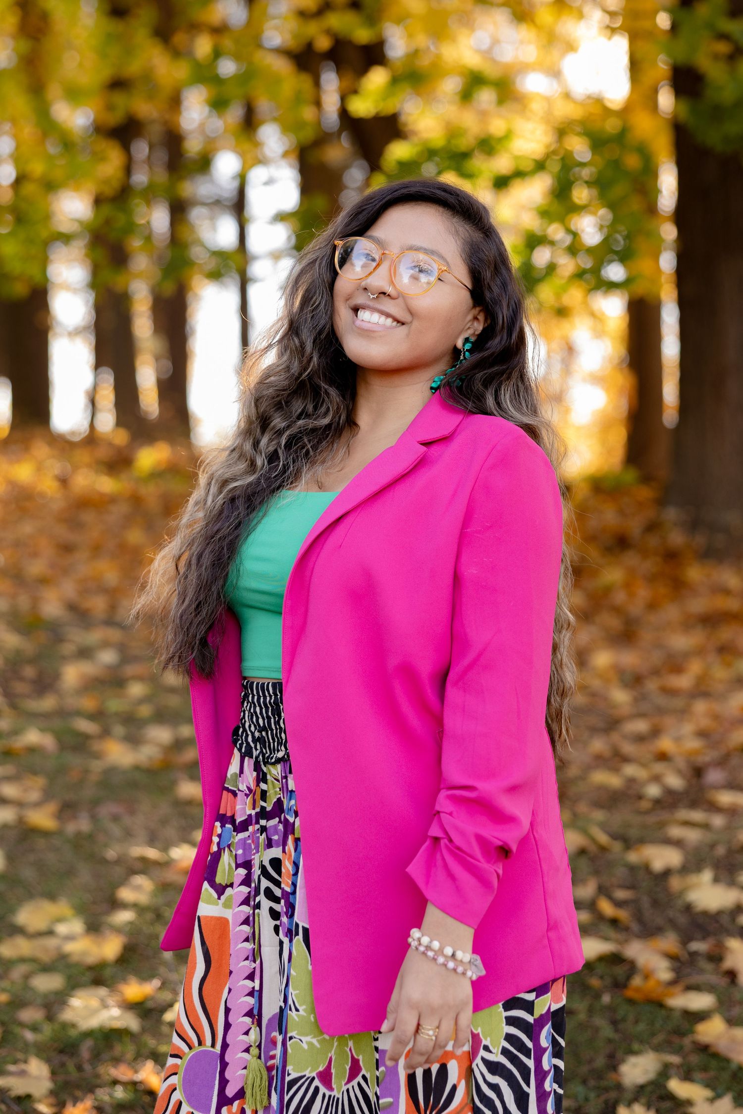 Joyful person wearing hot pink blazer and patterned dress poses in autumn forest setting with golden leaves.