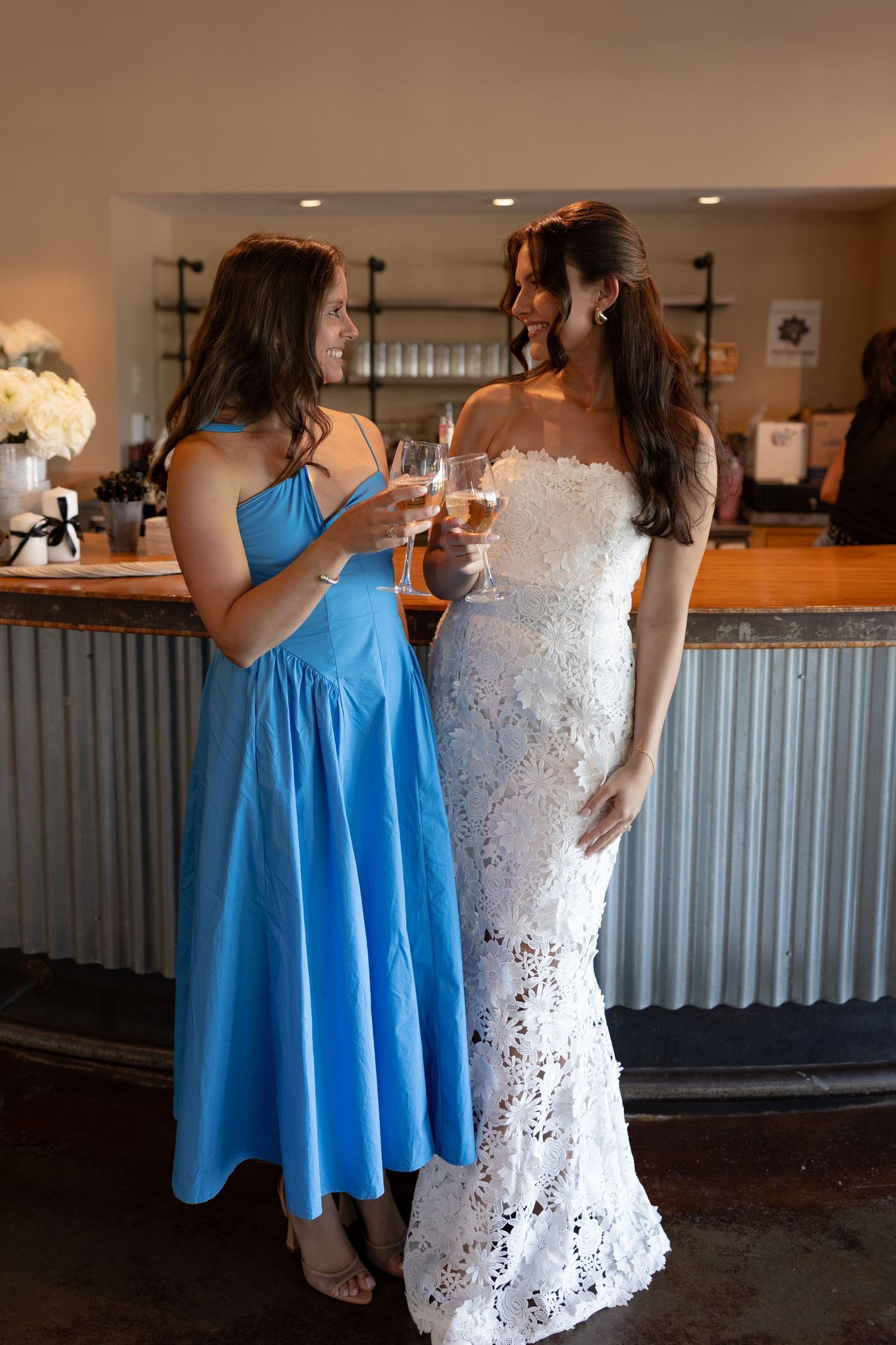 Wedding party members in blue dresses and white gown share drinks at a rustic indoor bar.
