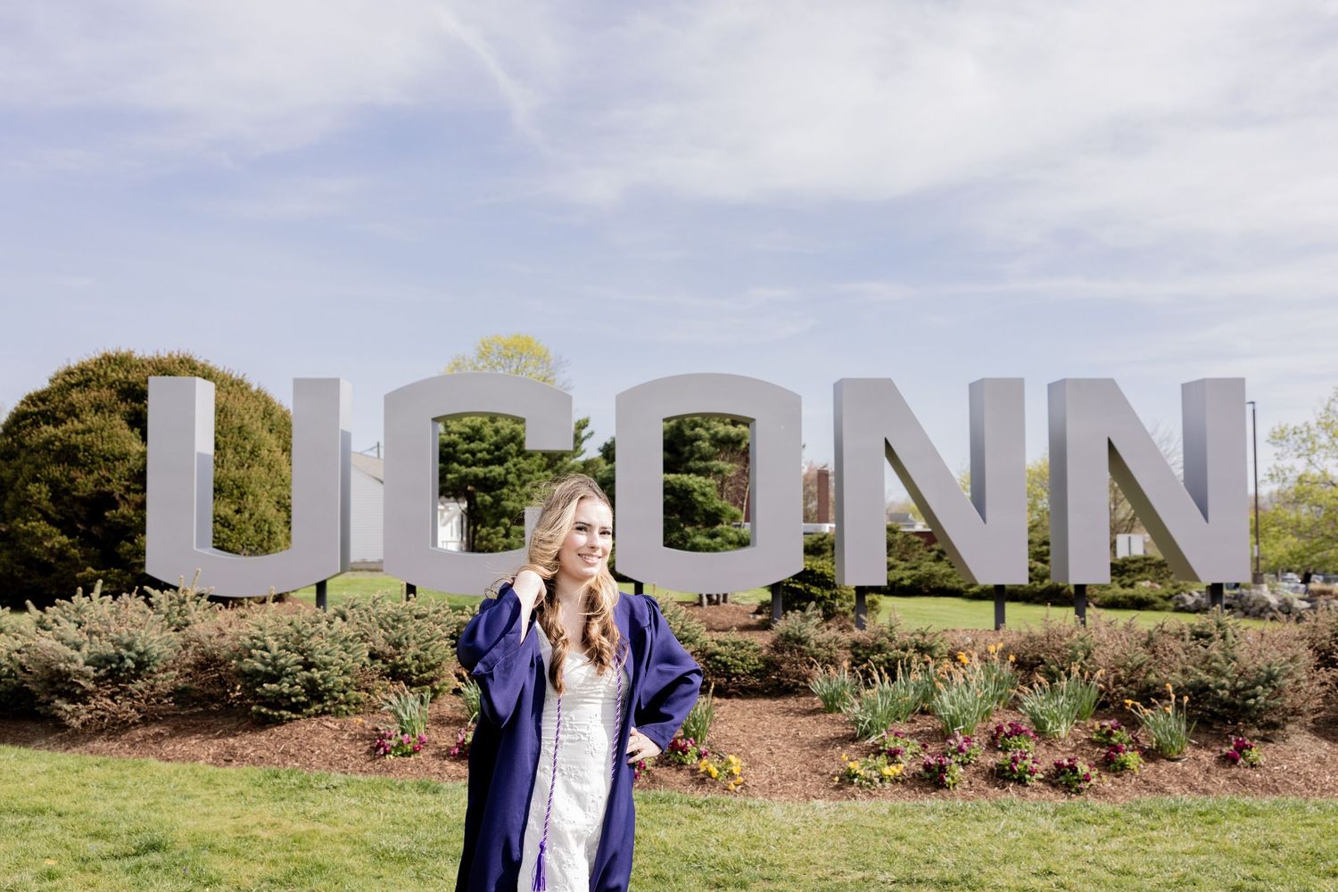 Graduate stands in front of large UCONN letters sign surrounded by landscaped gardens on a sunny day.