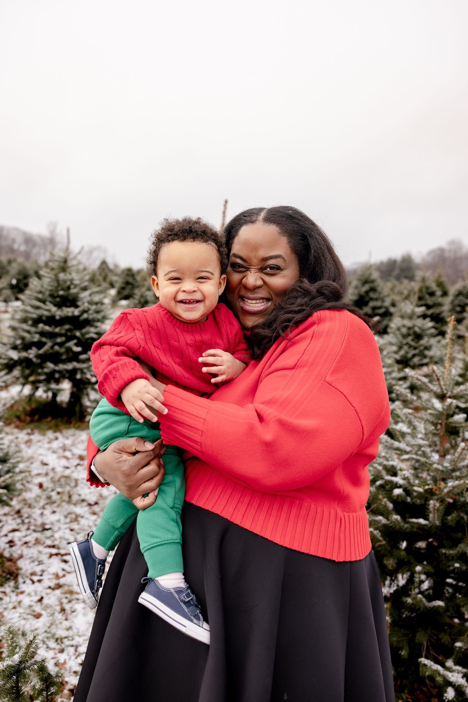 Two people in red and black clothing share joyful embraces at a snowy Christmas tree farm.