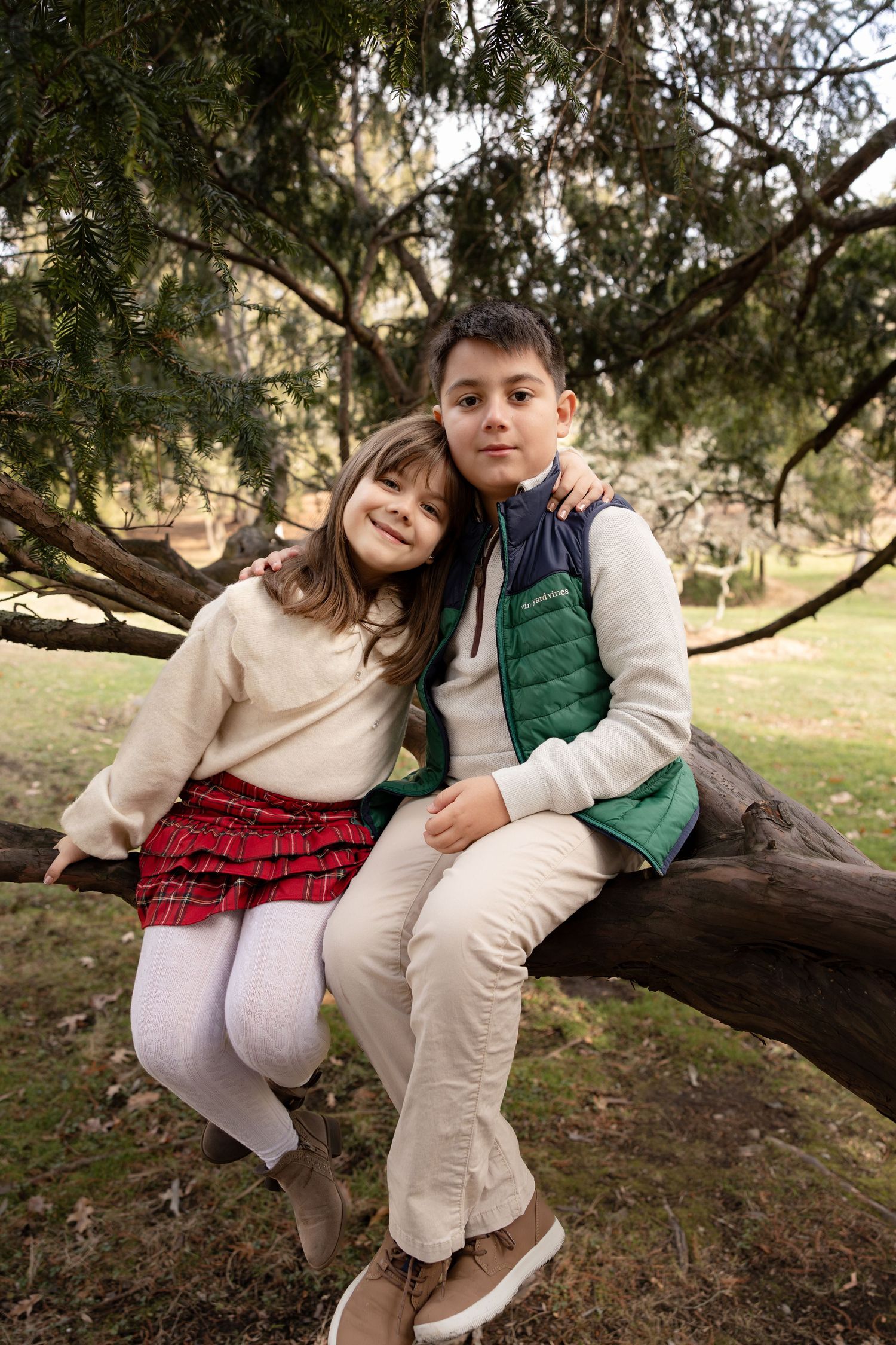 Two children sit and play together on a fallen tree branch in a park setting during autumn.