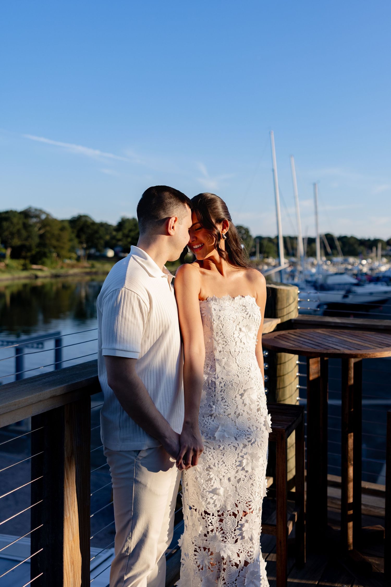 Romantic couple poses on a waterfront dock with boats and marina in the background at sunset.