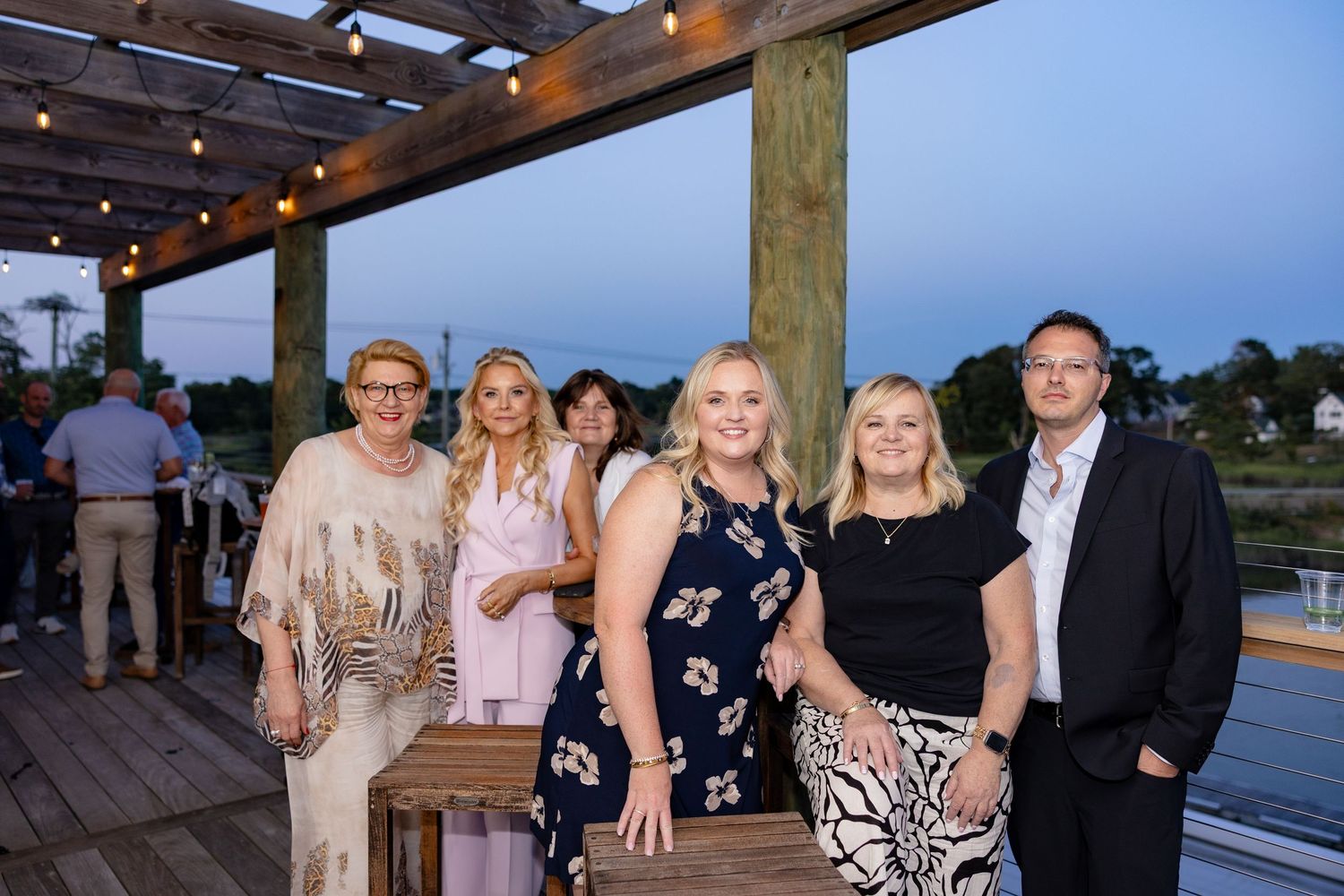 Wedding guests pose for outdoor photos at sunset on a wooden deck.