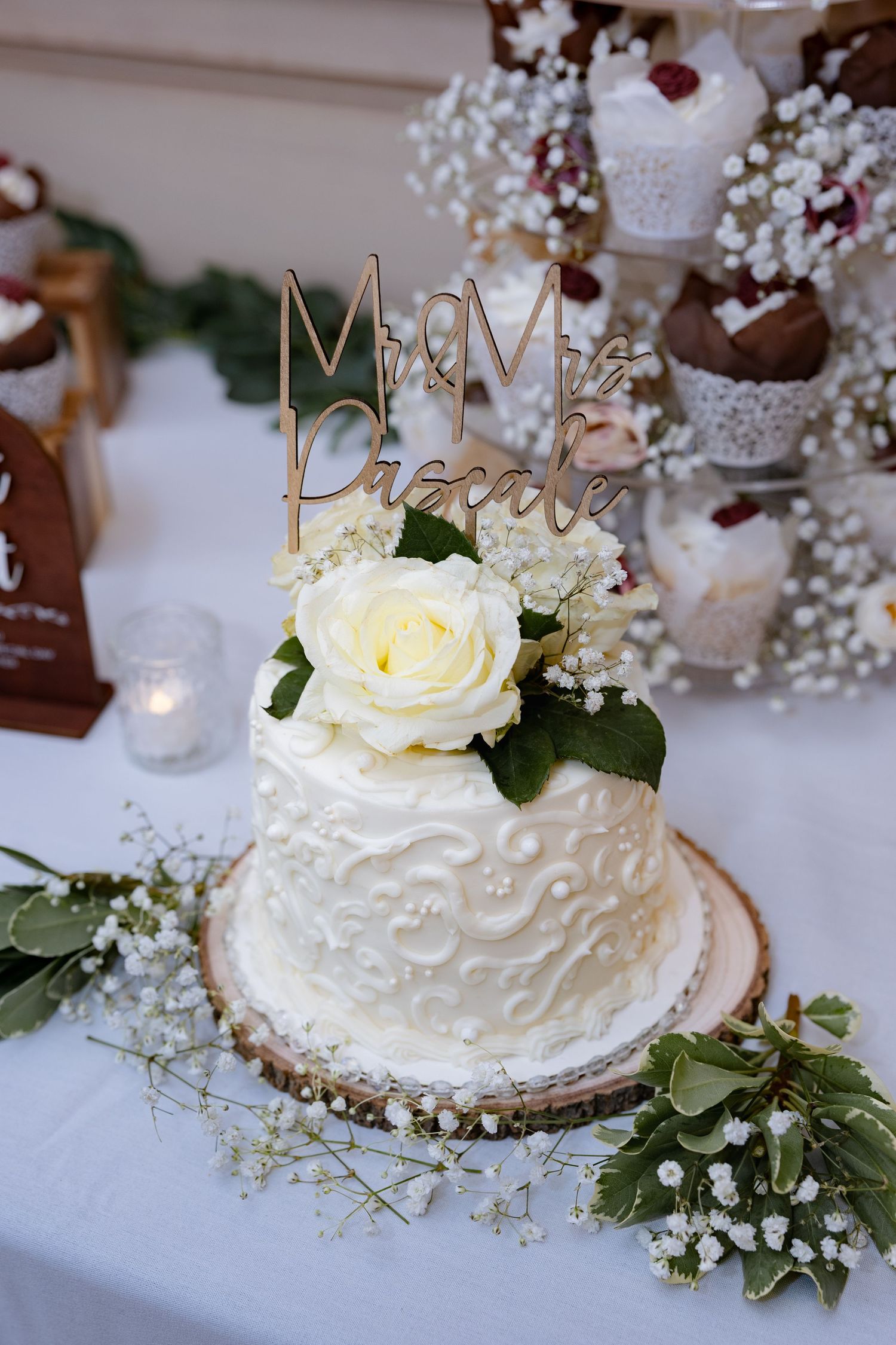 A white wedding cake decorated with roses and baby's breath sits on a rustic wooden base with a gold 'Mr & Mrs' topper.