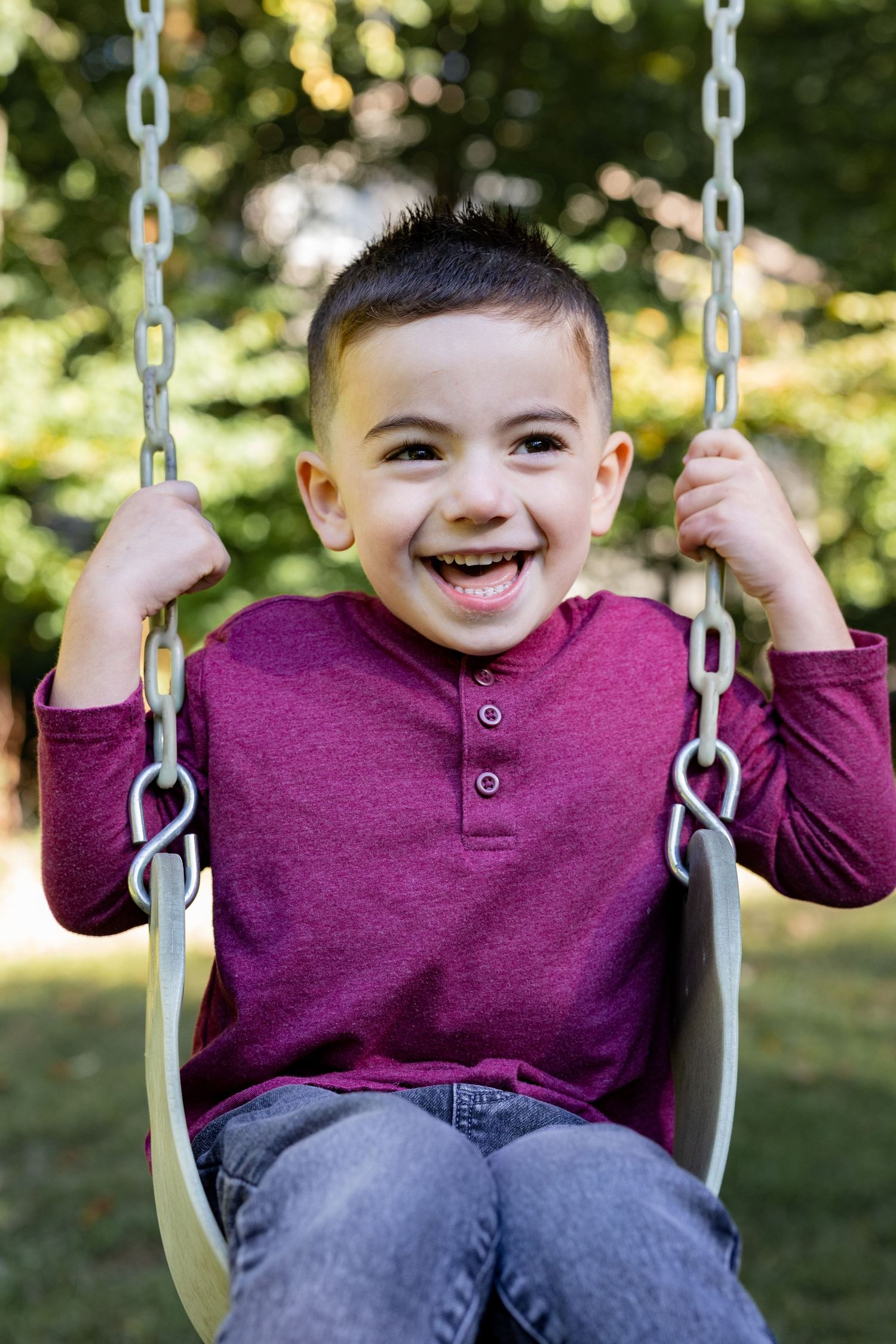 A joyful child wearing a burgundy henley shirt swings outdoors on a sunny day with trees in the background.
