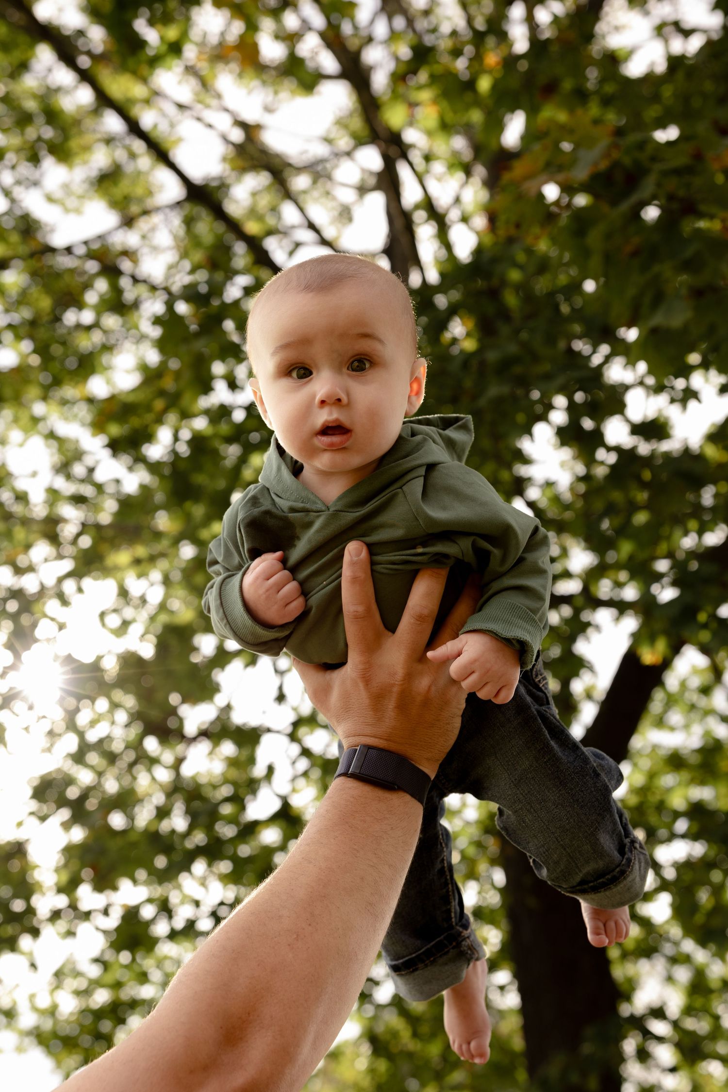A baby wearing a green hoodie is lifted up against a backdrop of sunlit trees and foliage.
