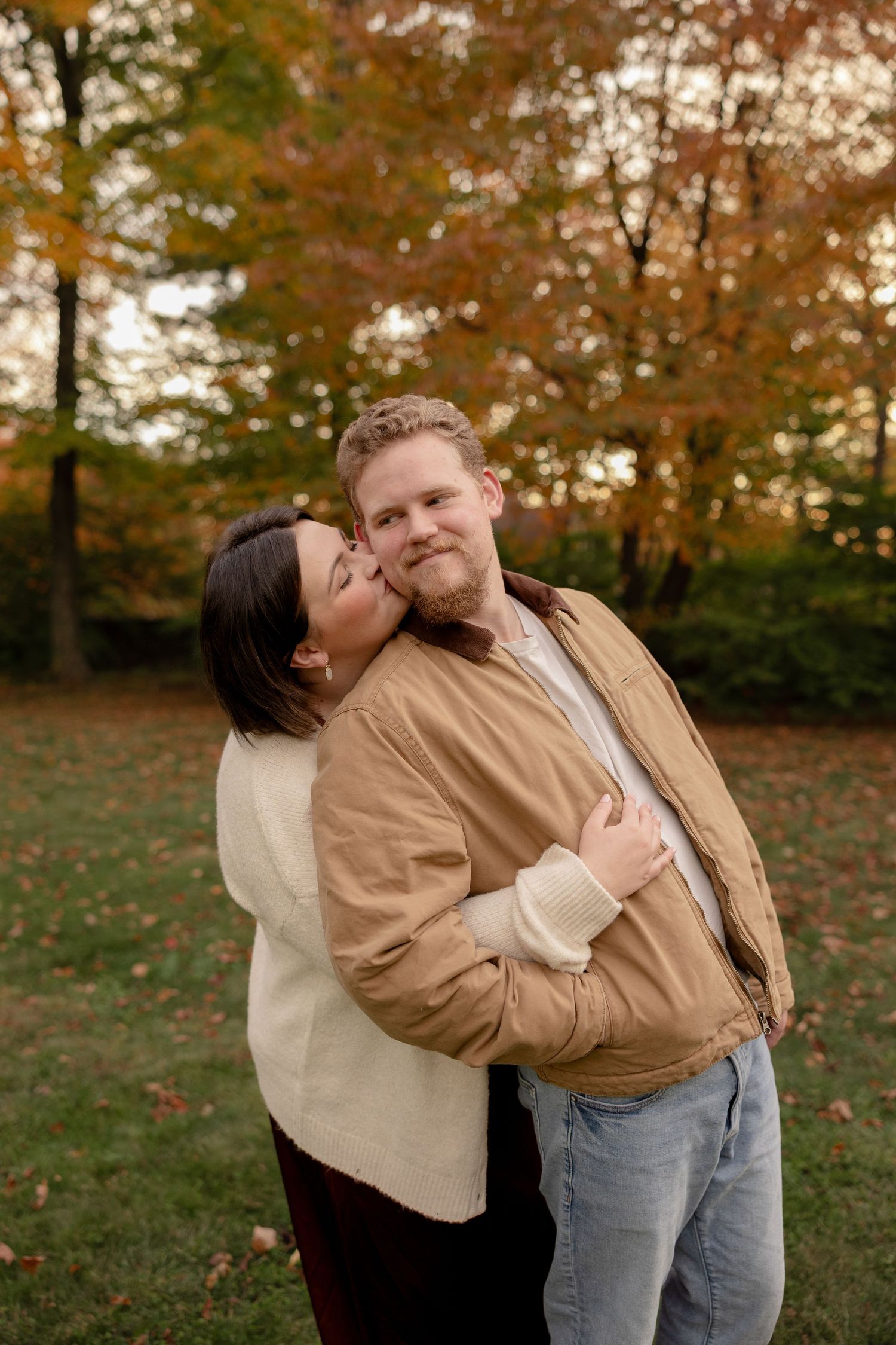 A couple embraces while standing in a park with autumn foliage in the background during golden hour.