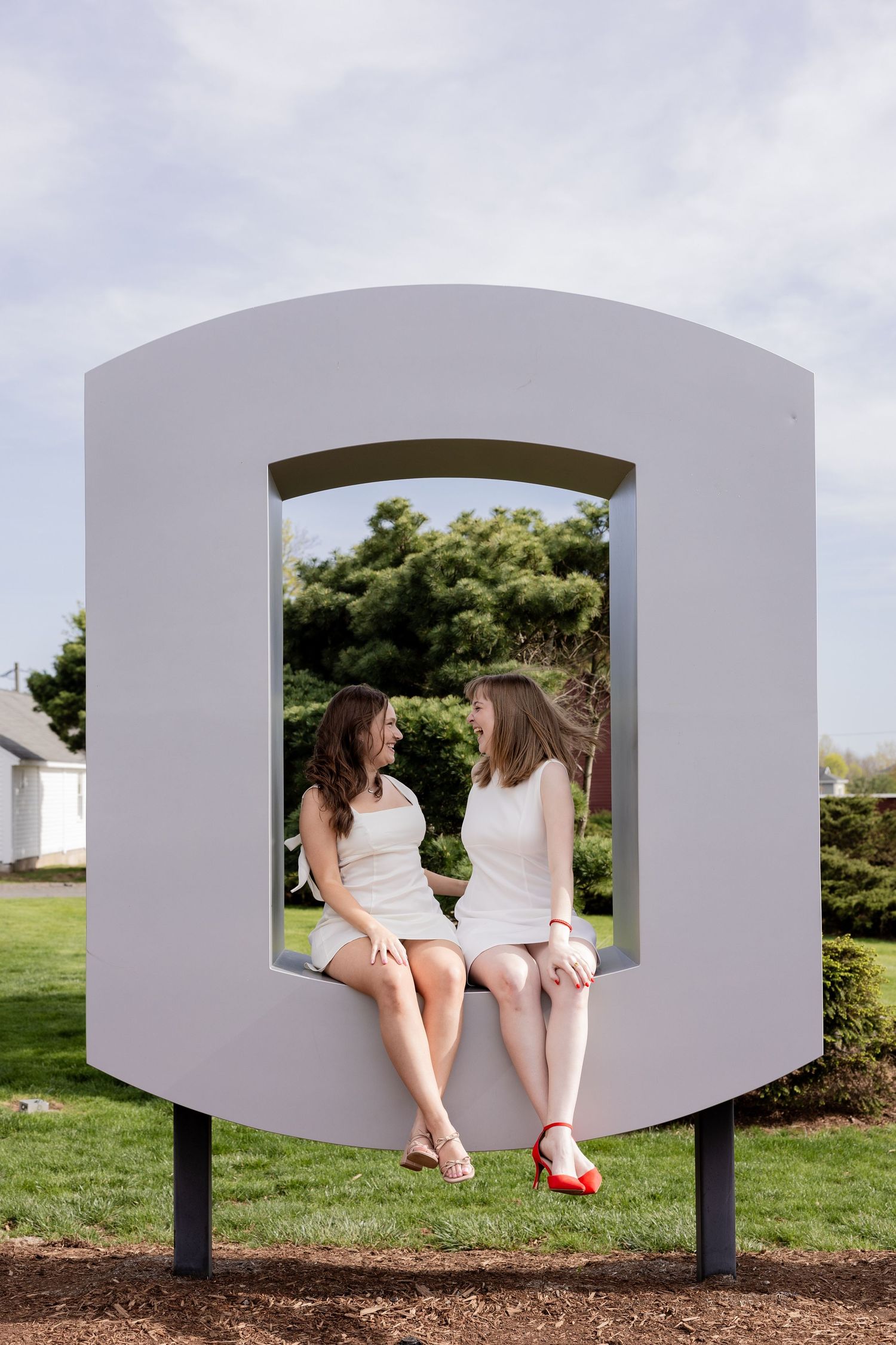 Two figures in white outfits sit together in a large artistic white frame installation on a grassy lawn.