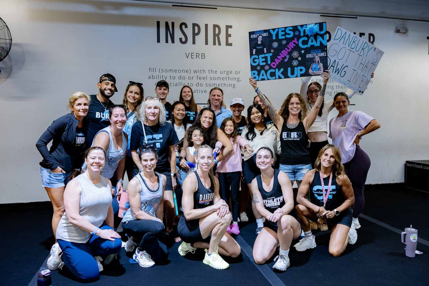 Group fitness class participants gather for a celebration photo beneath an 'INSPIRE' sign at a boutique gym.