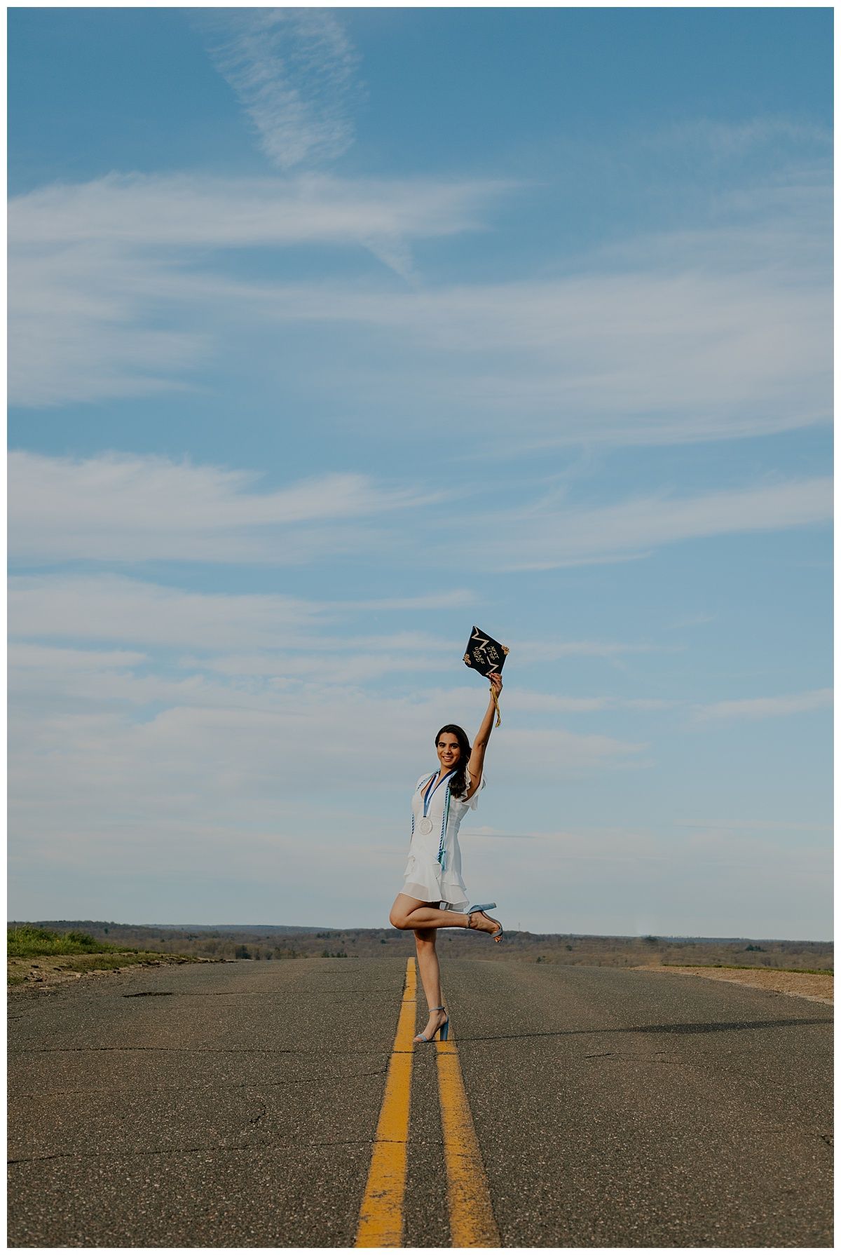 Graduate in light dress stands on yellow road line holding cap against blue sky.