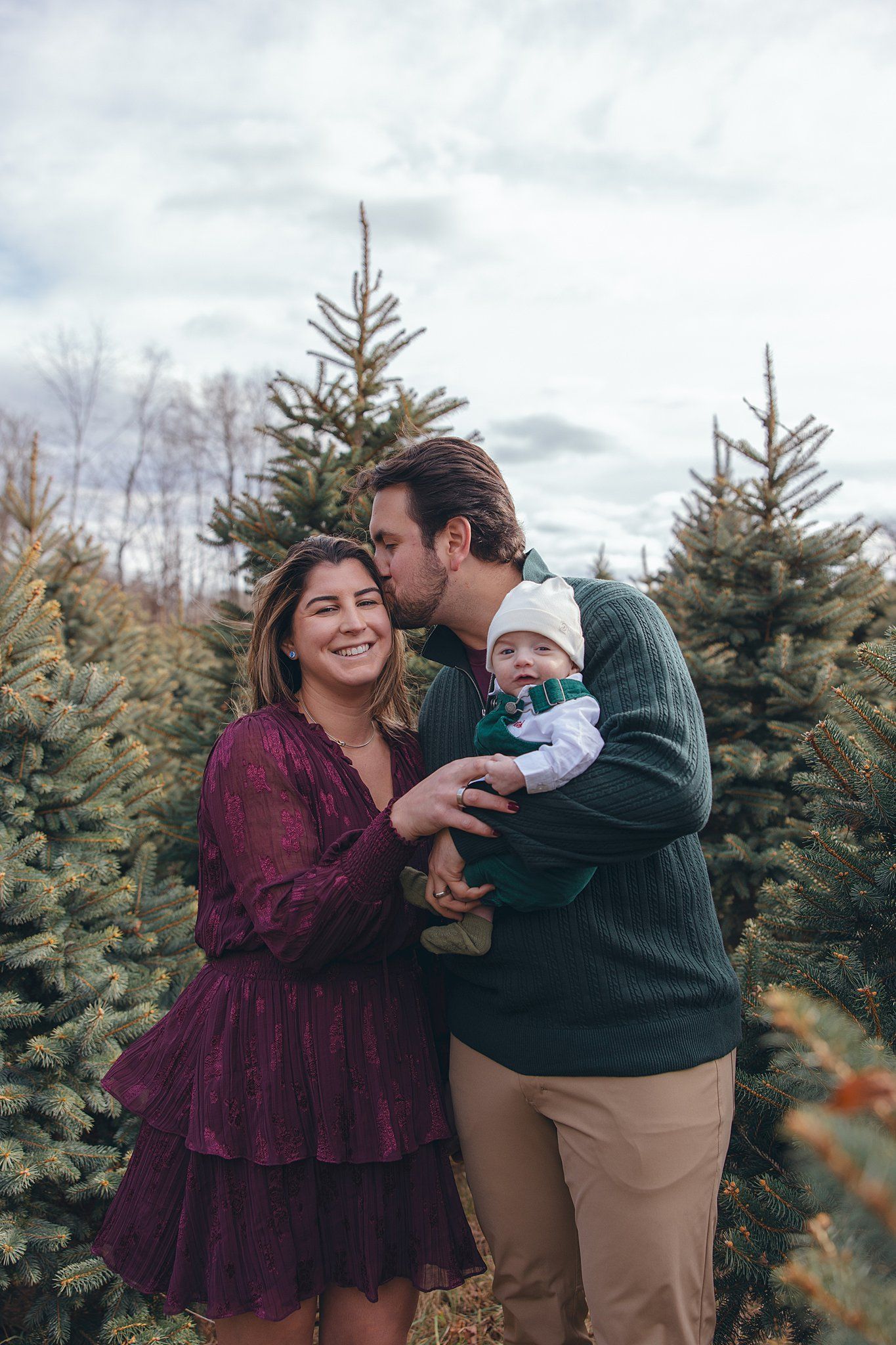 mom and baby smiling at camera and dad kissing mom on the cheek in front of a pine tree on maple row farm for holiday photo