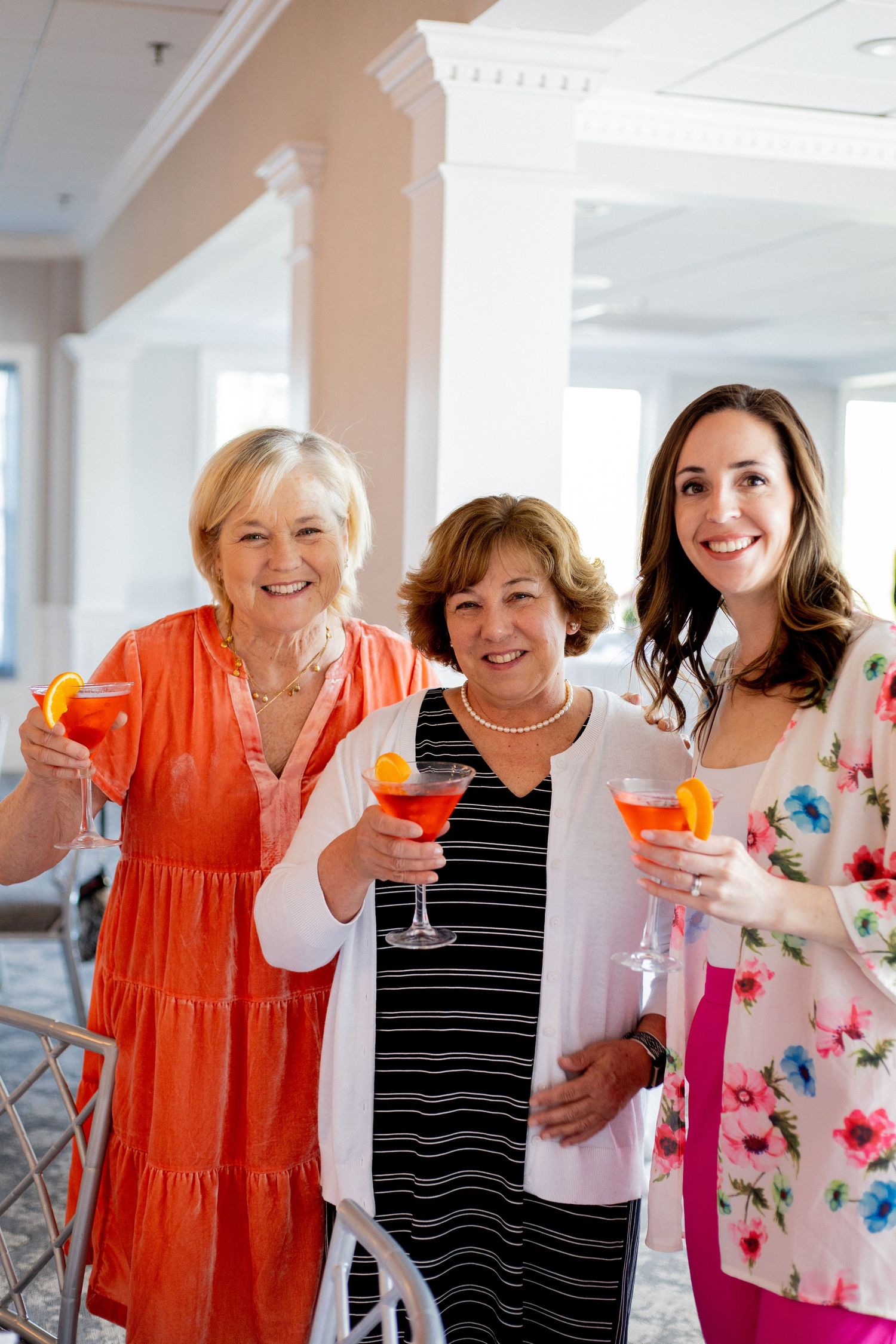 Three women hold orange cocktails while celebrating at an indoor event.