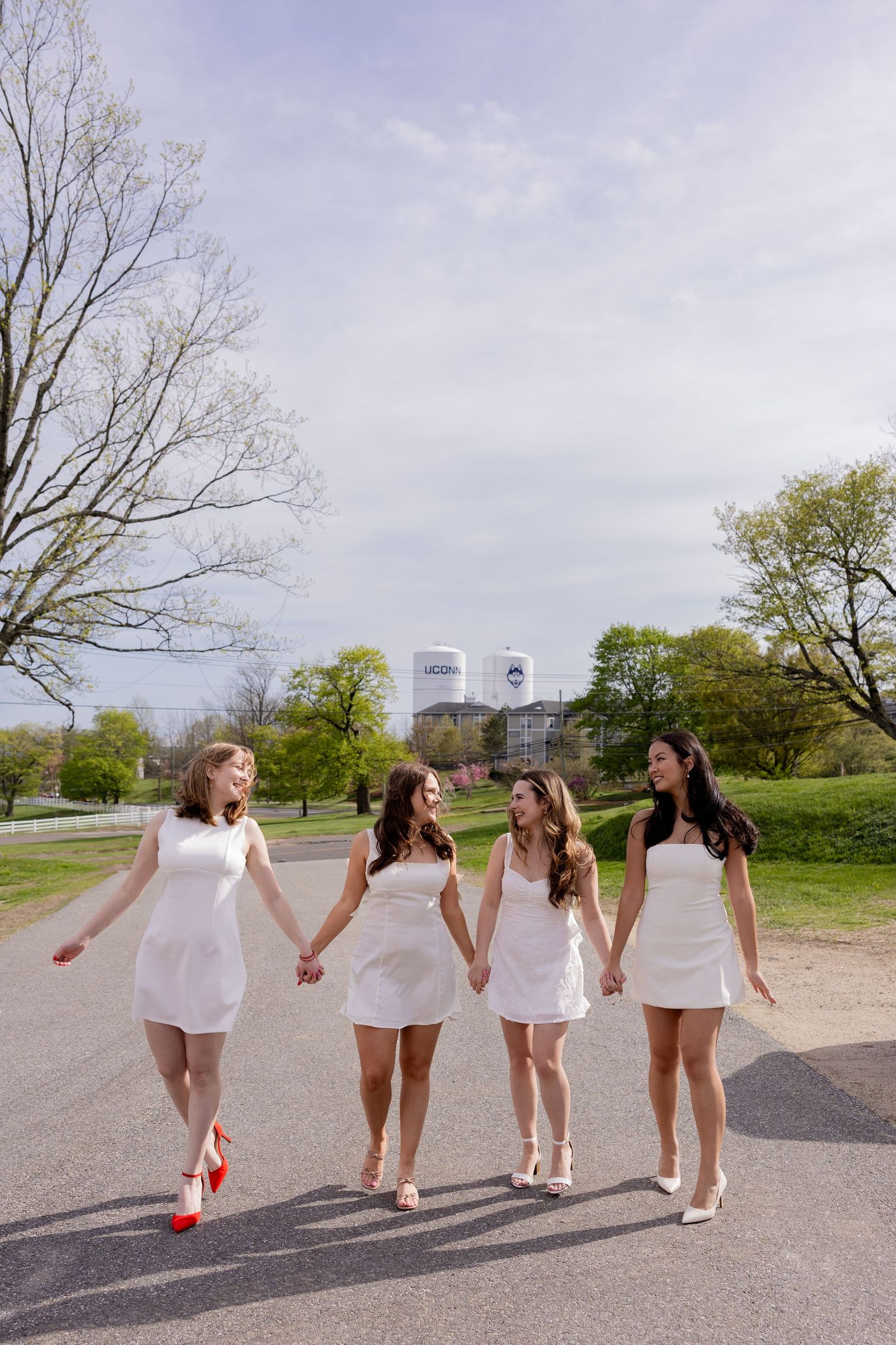 Four friends in matching white dresses walk together on a path with city buildings visible in the background on a spring day.