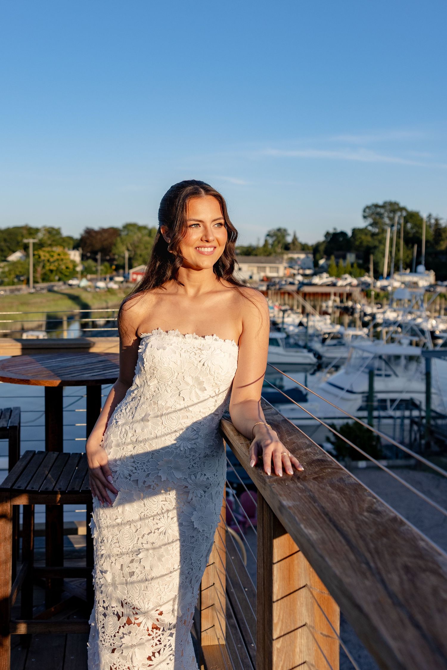 A woman in a white lace dress poses casually on a wooden deck with a marina and boats in the background.