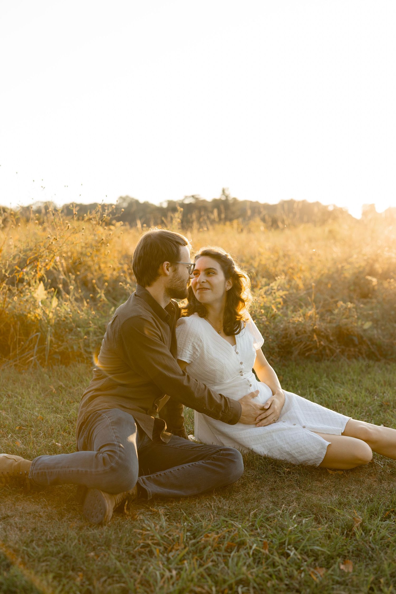 A couple lies together on a blanket in a grassy field during sunset, sharing an intimate moment.