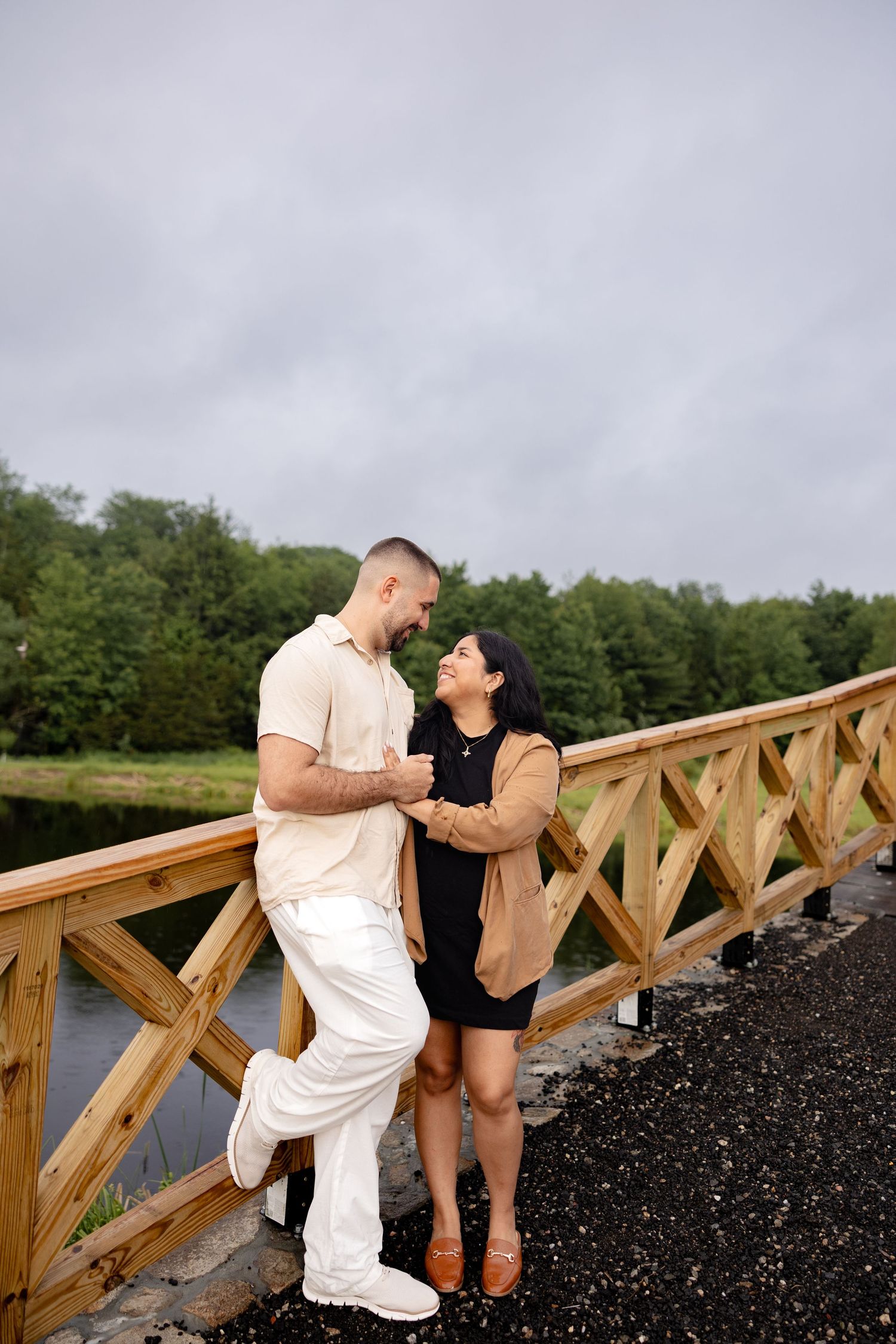 Two people share a romantic moment on a wooden bridge overlooking a scenic waterway on a cloudy day.