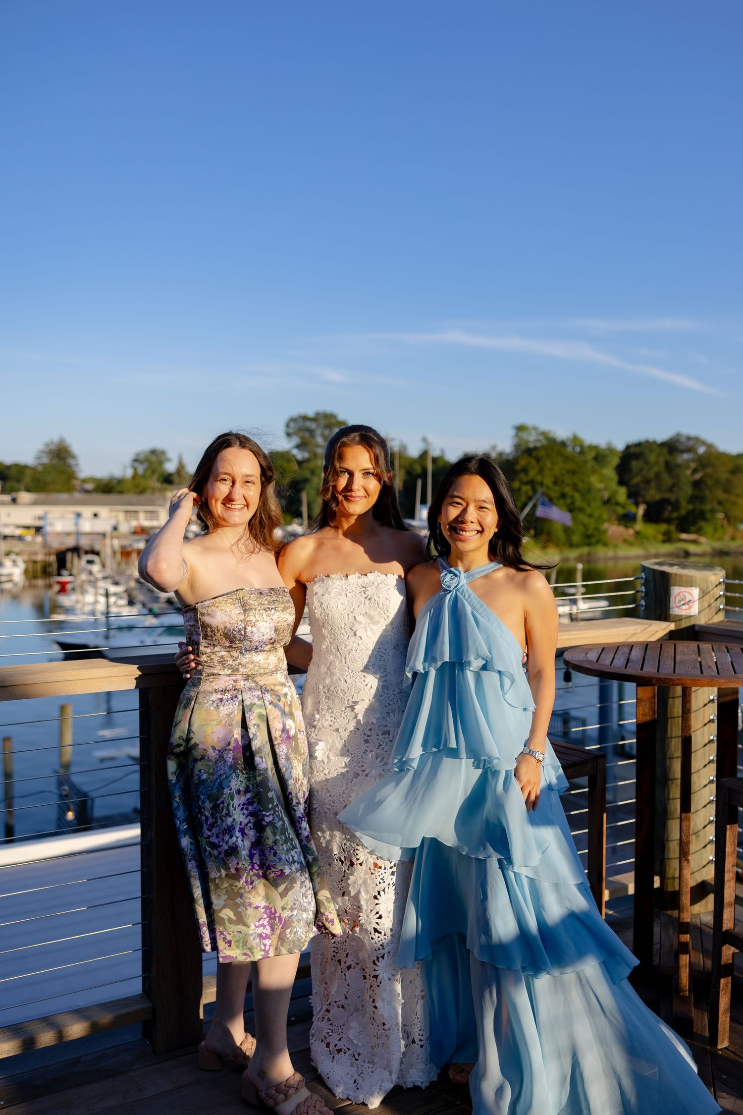 A group poses cheerfully on a waterfront deck wearing formal dresses in white, blue and floral patterns.