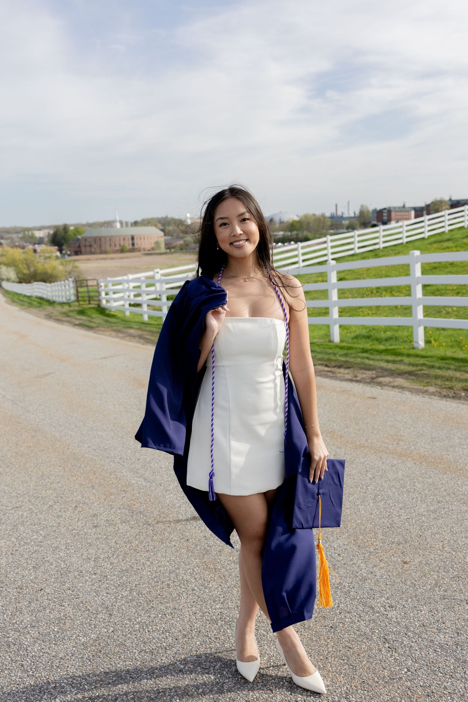A person in a white dress and navy graduation gown poses on a country road with white fences on a sunny day.
