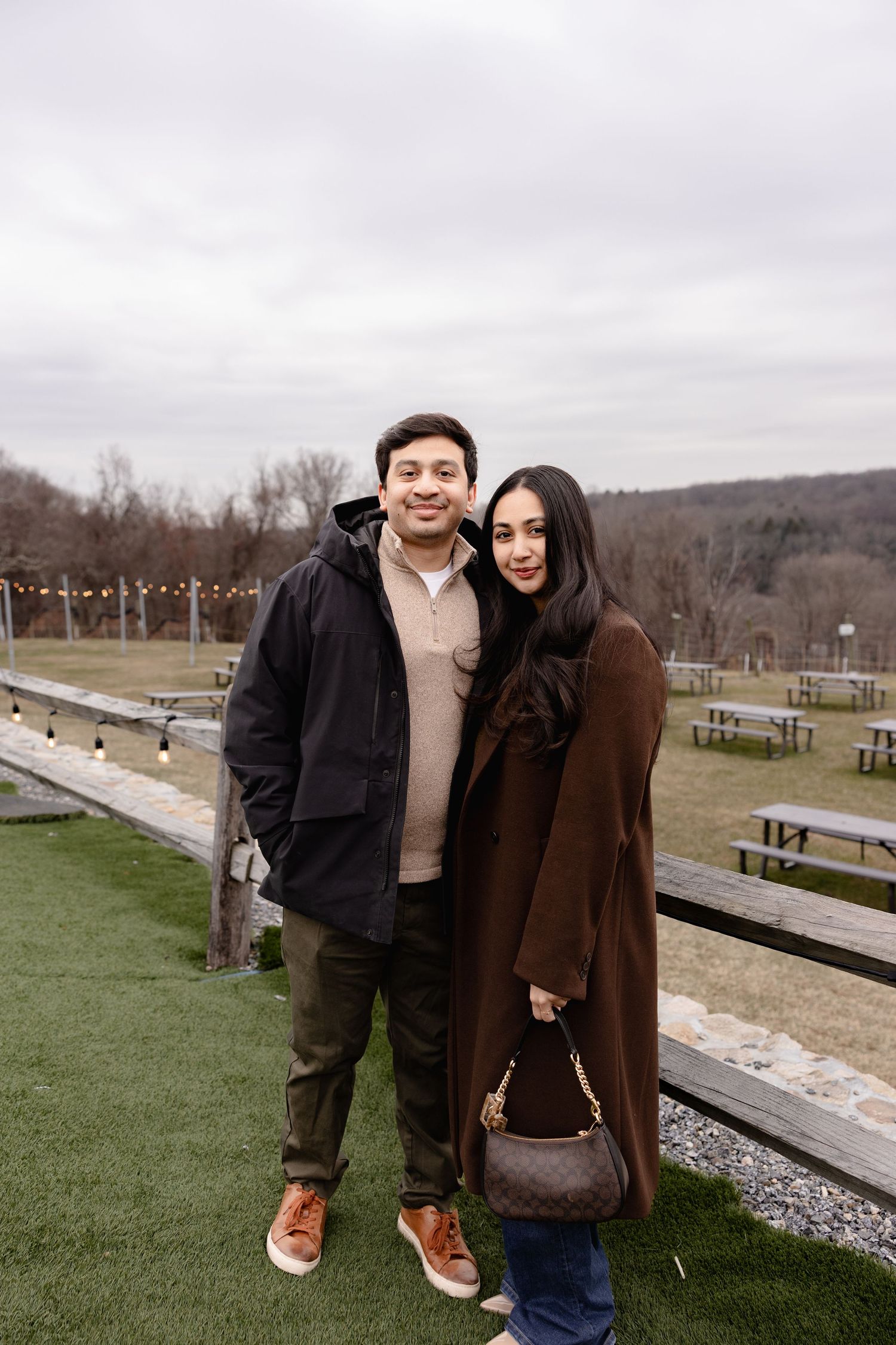 Two people in winter coats pose together on a cloudy day at a scenic overlook with hills in the background.